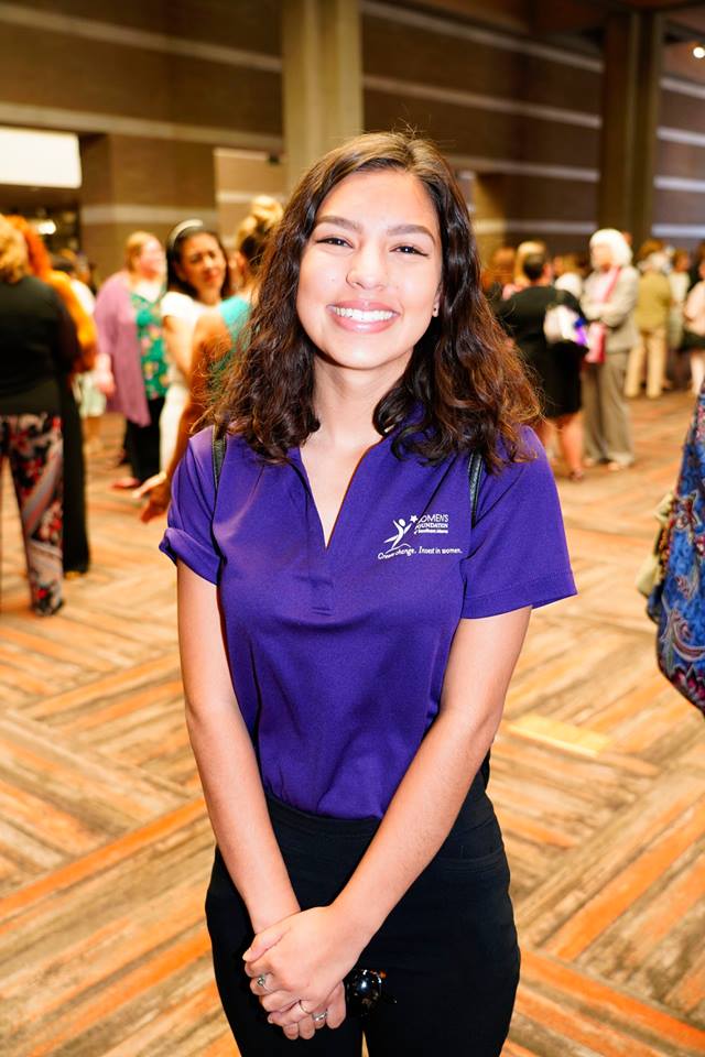 Single member of Unidas stands in a room, smiling for photo. Other Women's Foundation of Southern Arizona stand out of focus in the background.