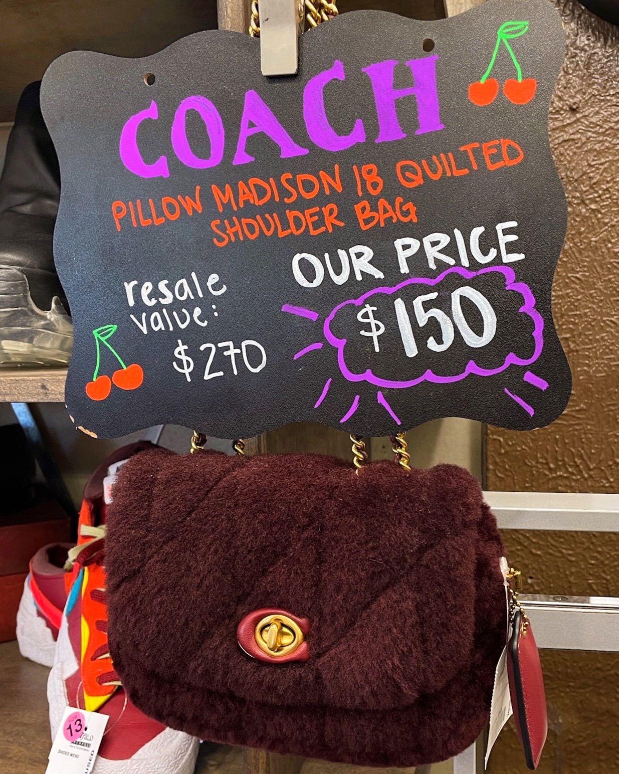 Sign above a handbag reads "Coach Pillow Madison 18 Quilted Shoulder Bag Resale Value $270 Our Price $150"