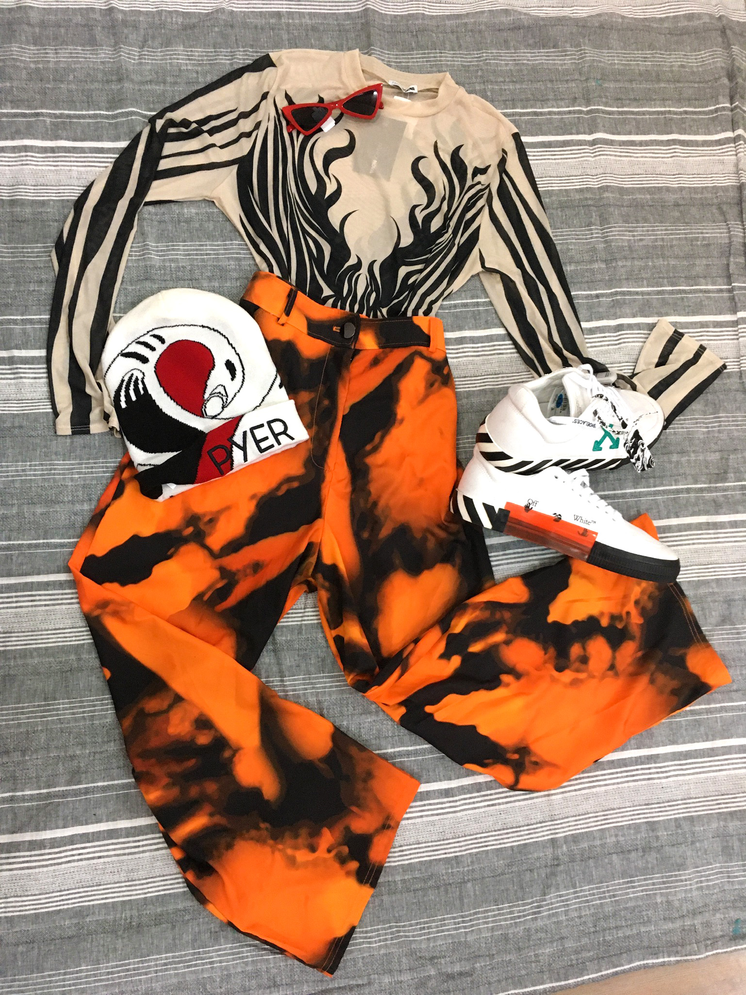 clothing laid flat, mesh longs sleeve top with swirling black pattern, orange and black tie-dyed pants, white sneakers and Pyer Moss logo beanie
