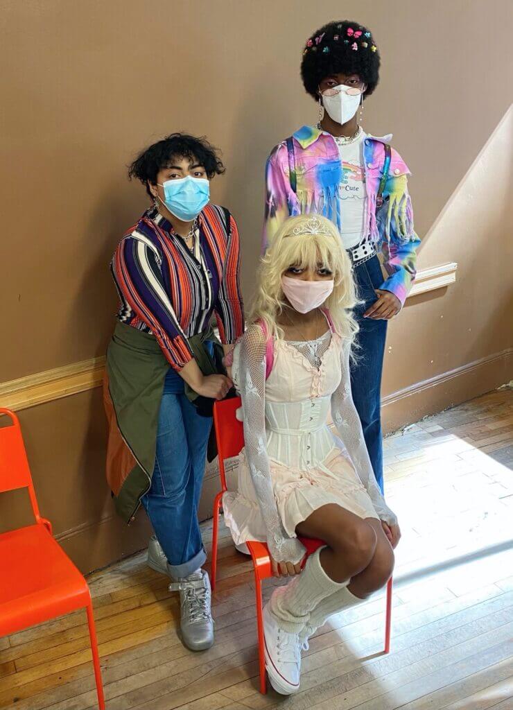 Two people wearing colorful shirts and denim, with another in a pink dress. All have accessories like silver jewelry, butterfly clips, and legwarmers.