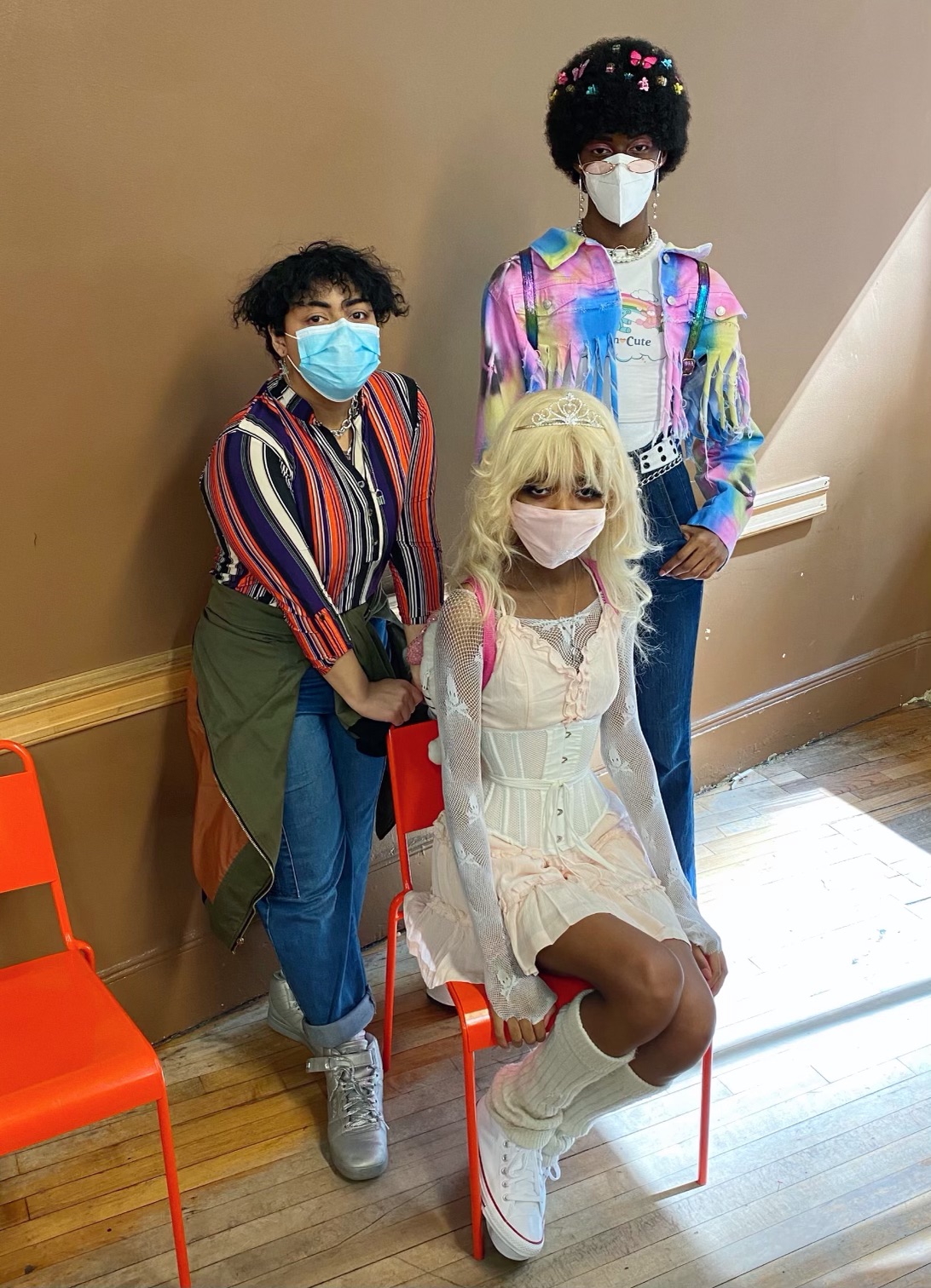 Two people wearing colorful shirts and denim, with another in a pink dress. All have accessories like silver jewelry, butterfly clips, and legwarmers.