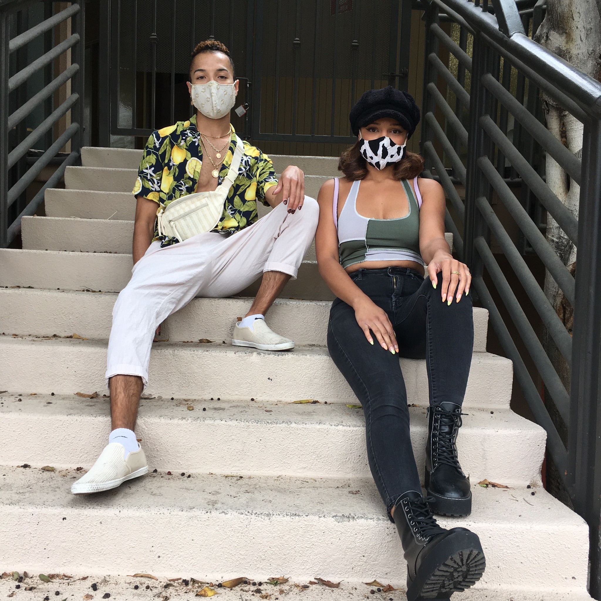 Two people on a cement staircase, one wears a lemon-patterned button down shirt and white pants, the other a patchwork tank and black denim.