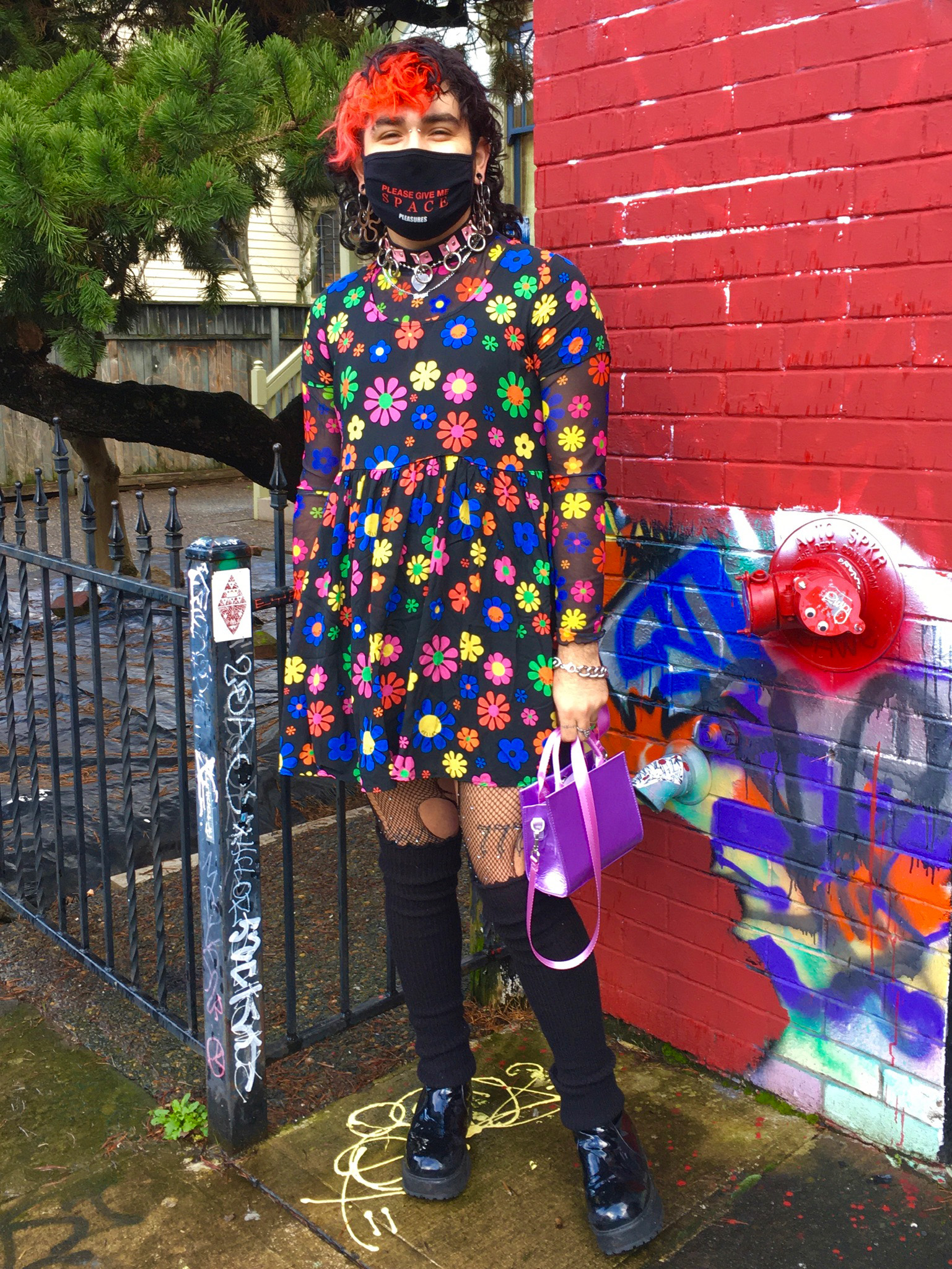 Person wearing black mesh dress with brightly colored flowers, distressed tights, purple satin handbag, and platform boots.