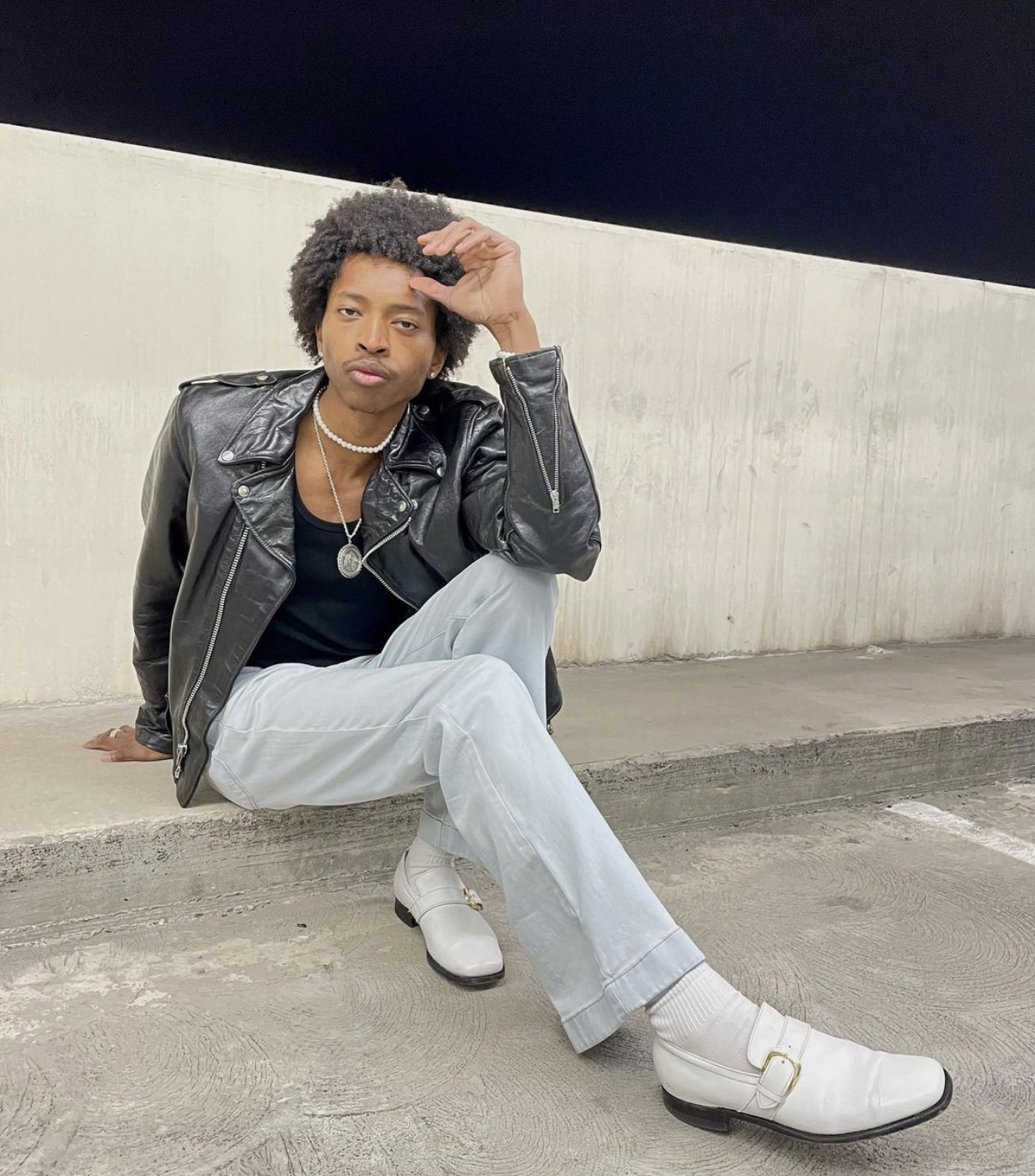 person sits on floor of a cement parking structure posed towards the camera. They are wearing a black leather motorcycle jacket, black t-shirt, light wash denim and white loafers with socks