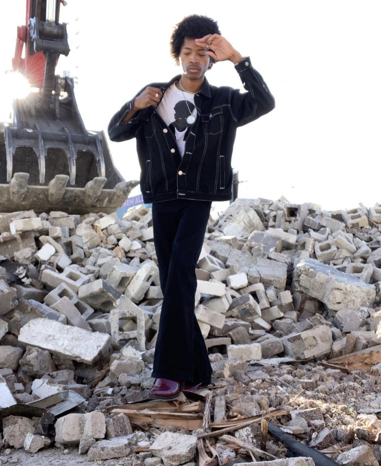 Katriel standing on a pile of cinder blocks wearing a black denim jacket over a white t-shirt, black flared pants and burgundy boots