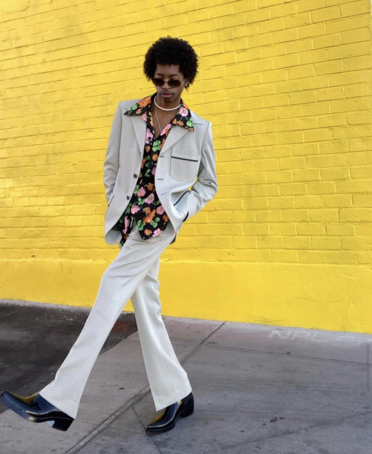a slim man poses in front of a bright yellow wall wearing a grey pinstripe leisure suit, floral button-down a black pointed-toe boots