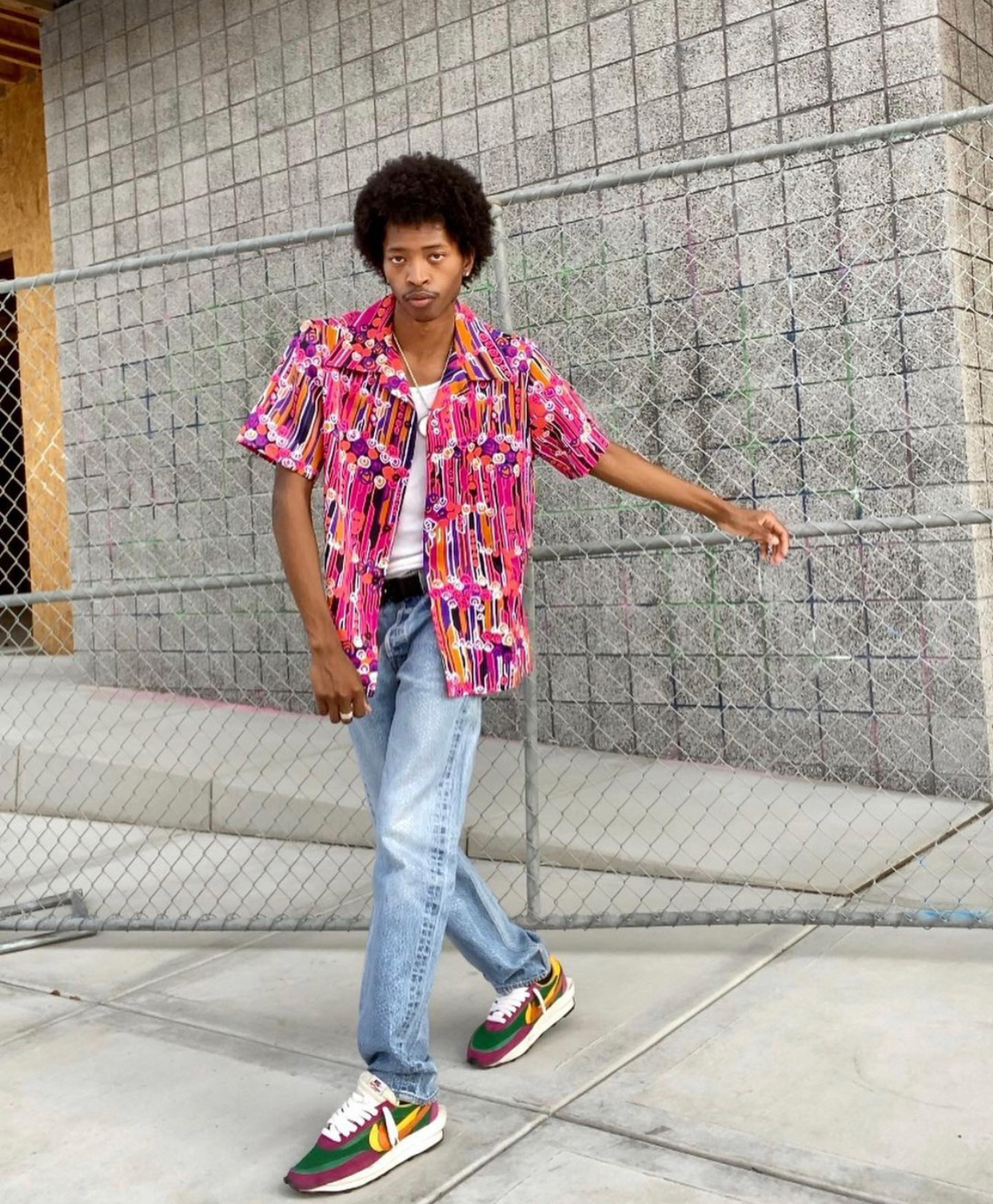 a man poses in front of a gray building and chainlink fence wearing a purple and pink abstract button-down with white t-shirt underneath, light wash denim and multicolored Nike sneakers