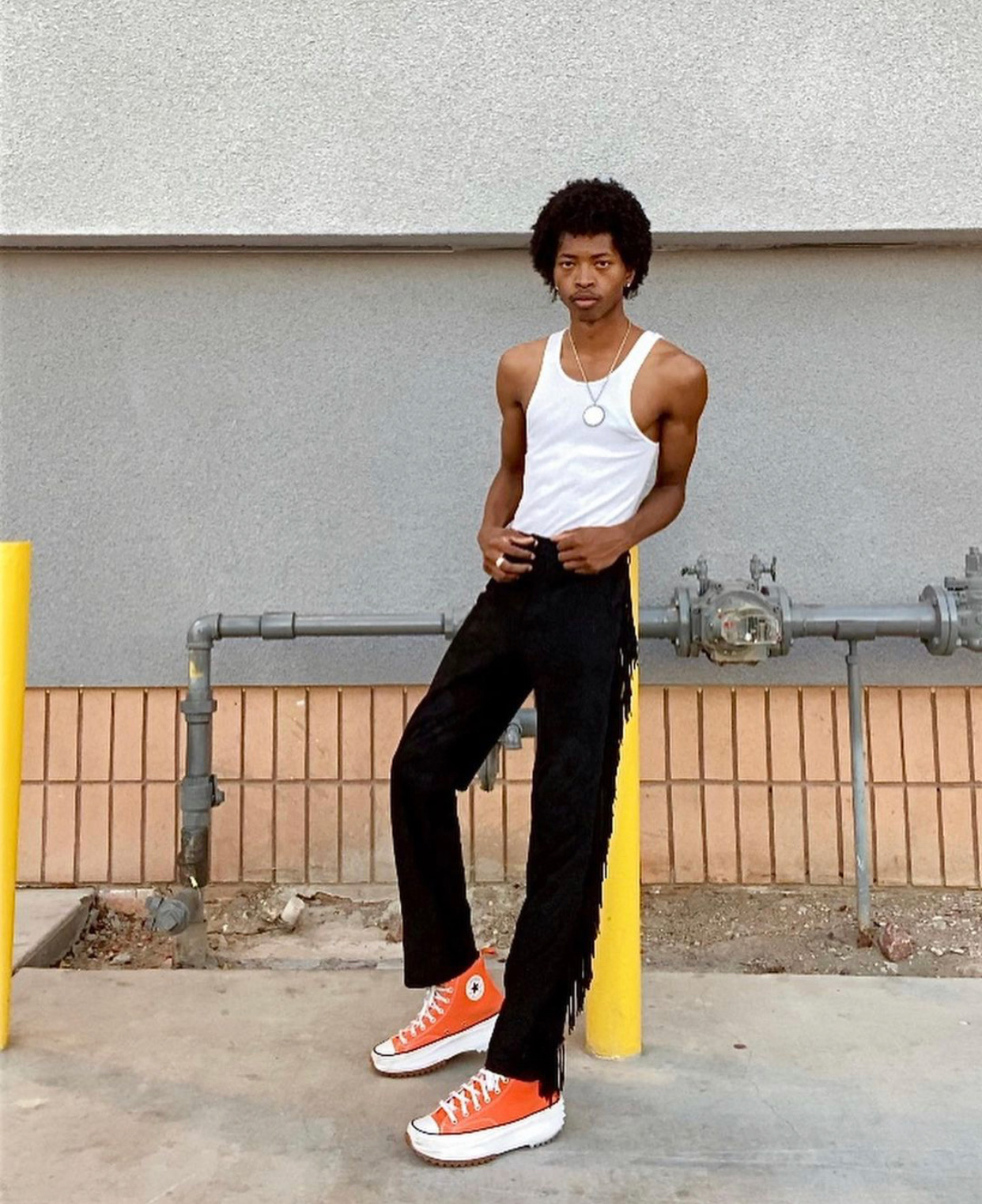 a slim man leans against a pole in a parking lot wearing a white tank top, black pants with fringe down the legs and orange platform Converse sneakers