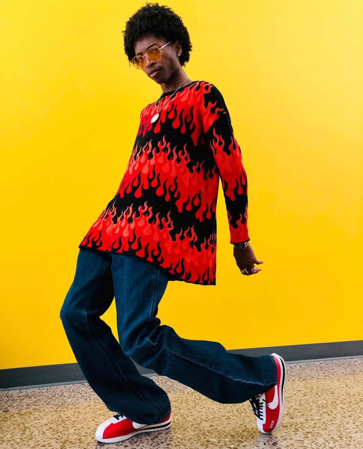 Person stands in front of a yellow wall wearing orange-tinted sunglasses, a flame-print sweater, baggy dark-wash jeans and red Nike Cortez sneakers
