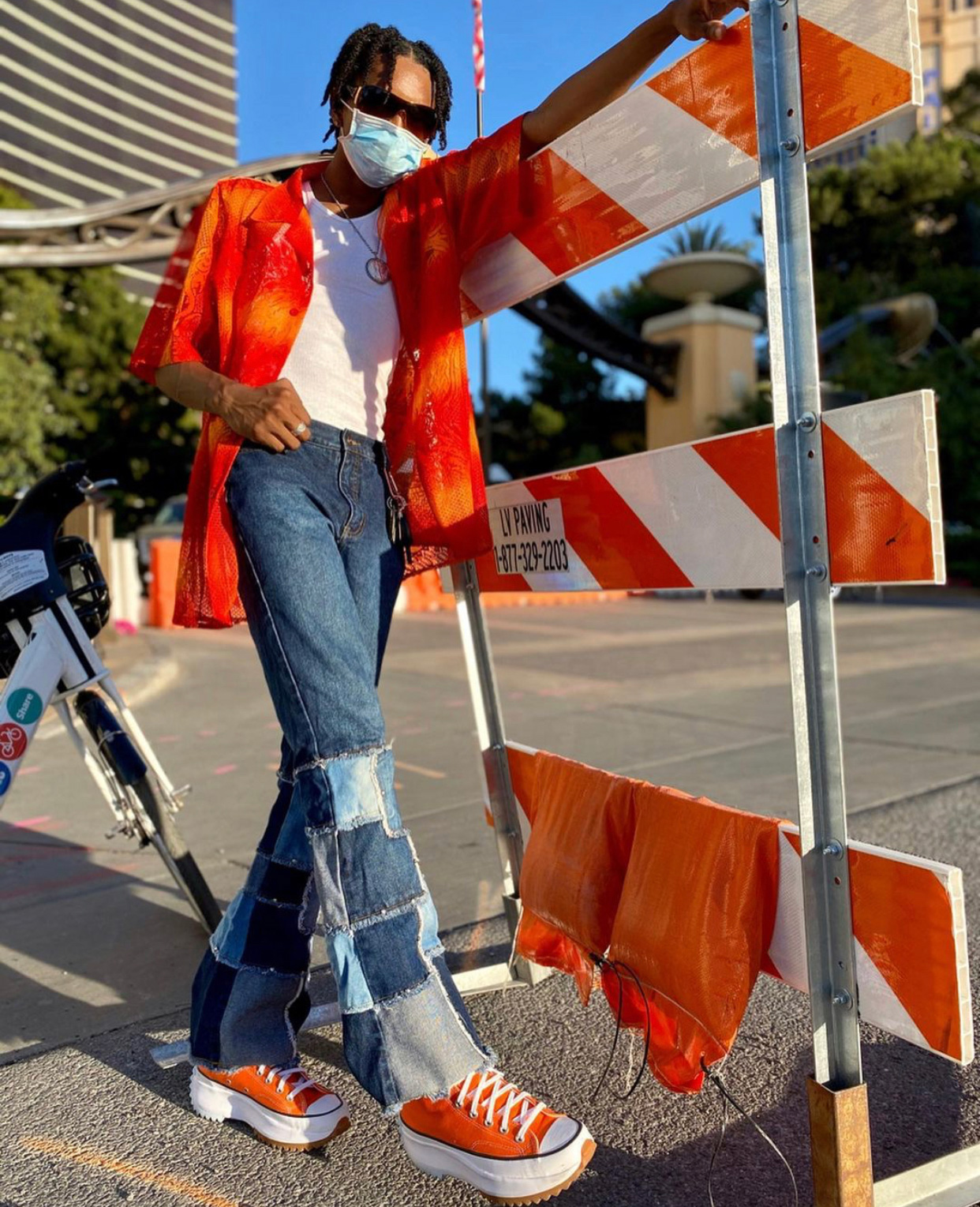 a slim man rests his arm on an orange traffic blockade wearing an orange and red abstract button down, white t-shirt, patchwork blue jeans and orange platform Converse sneakers