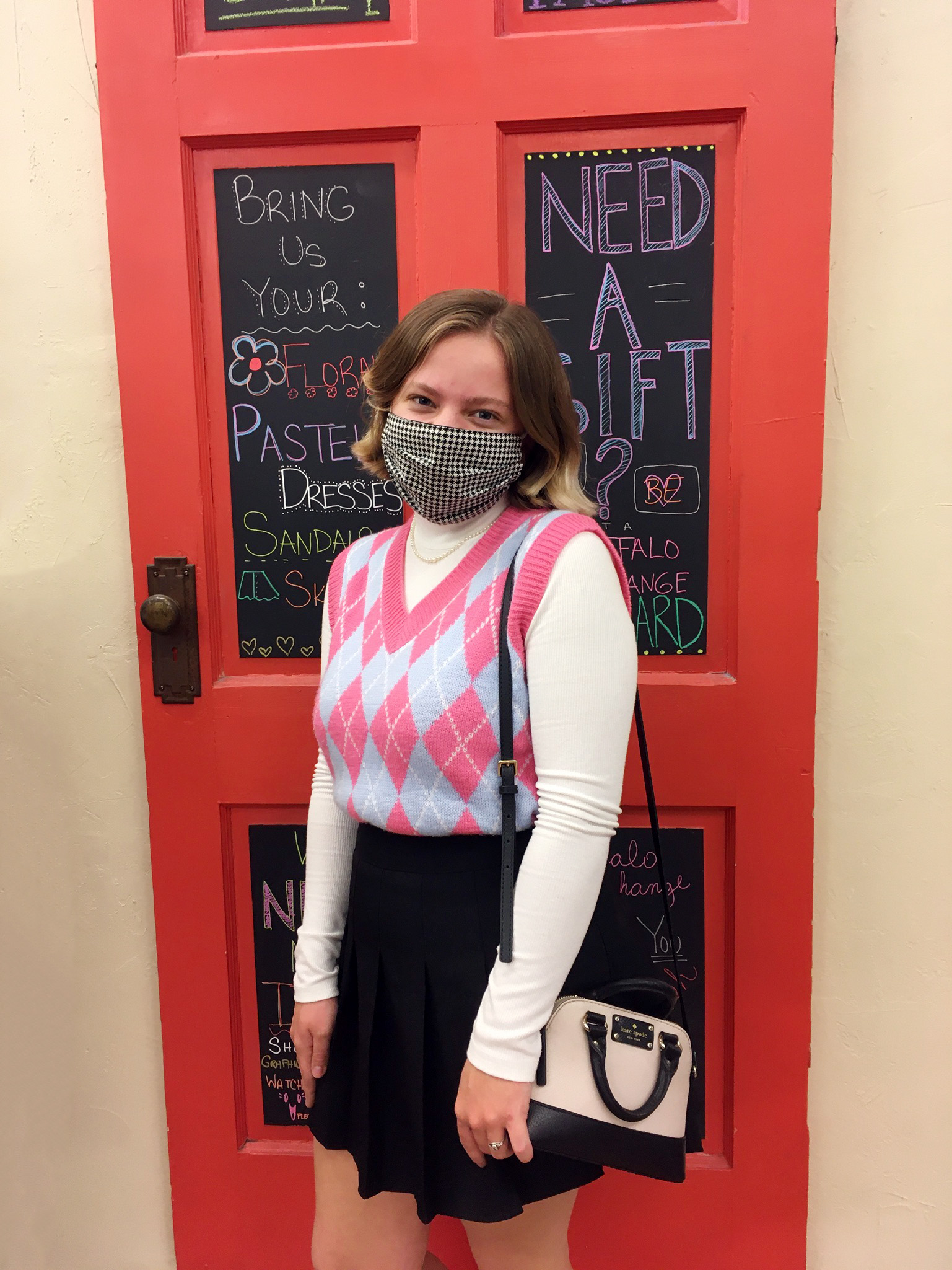 Person wearing face mask, argyle sweater vest styled with turtleneck underneath and pleated skirt with a Kate Spade crossbody bag on her shoulder