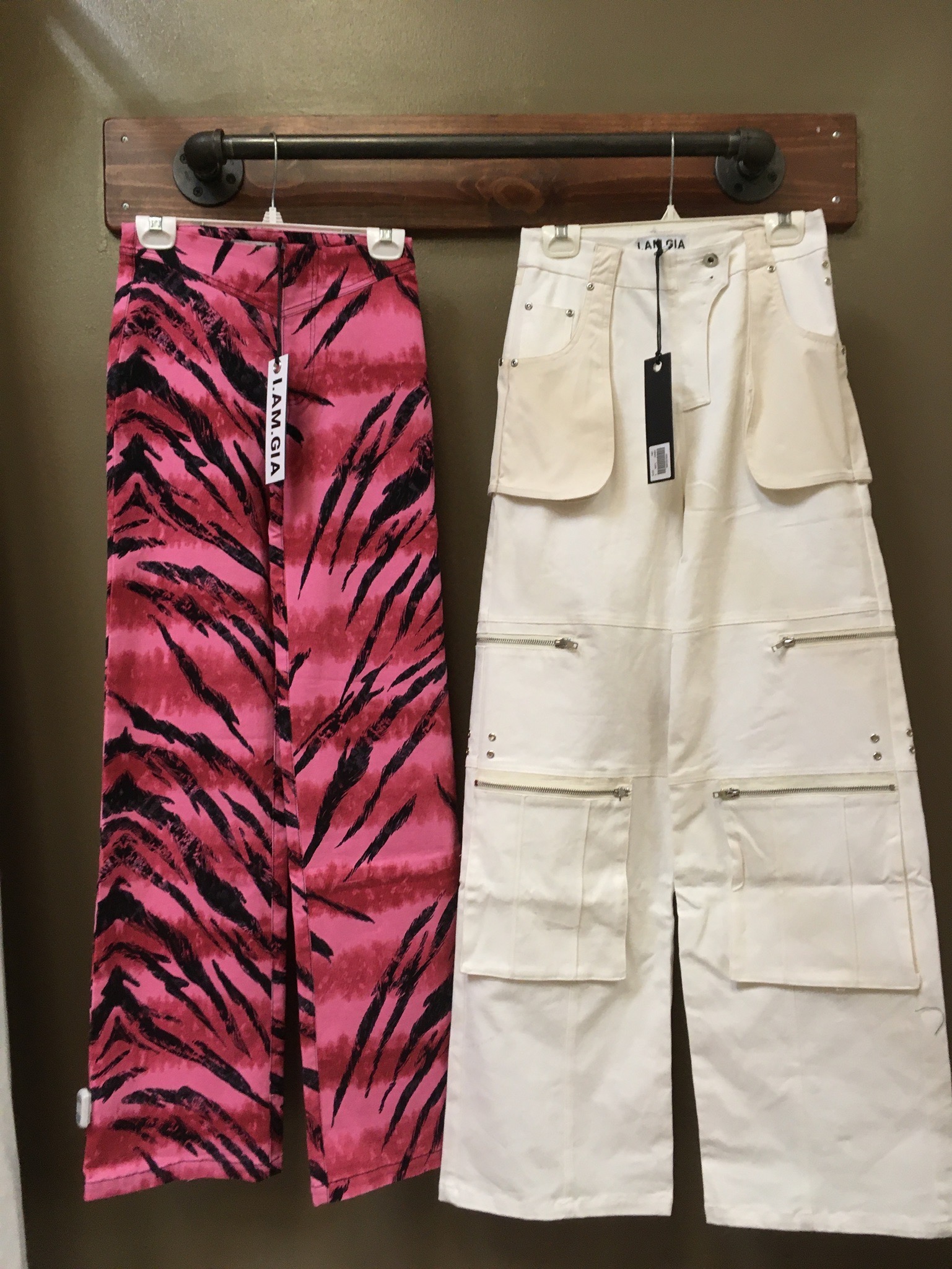 Two pairs of I.Am.Gia pants hang on a wall mounted garment bar. The left pair is pink tiger-print and the right are an off-white pair of cargo pants