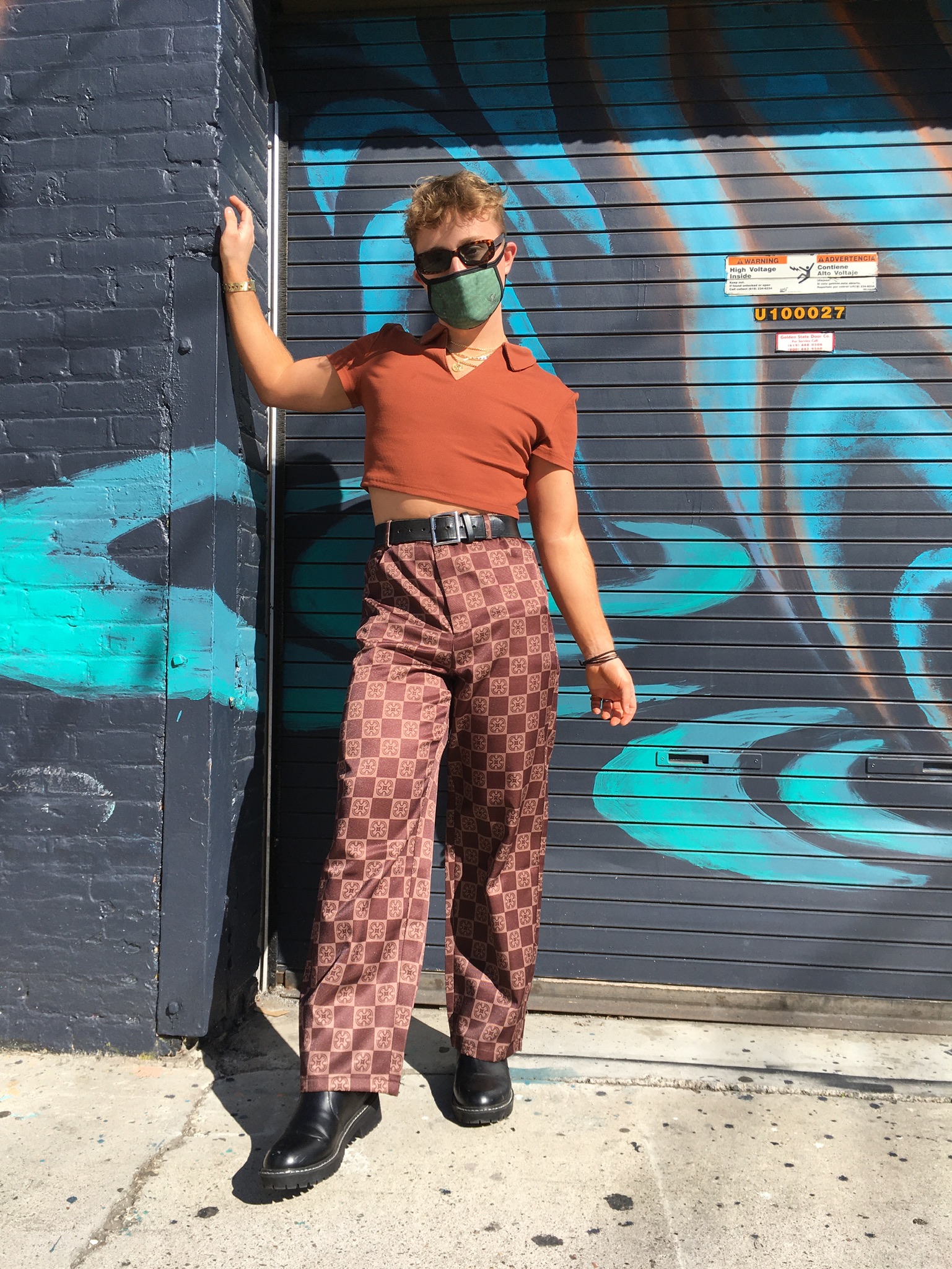 Person poses in front of painted wall mural wearing checkered pants styled with cropped brown polo, black sunglasses, belt and boots