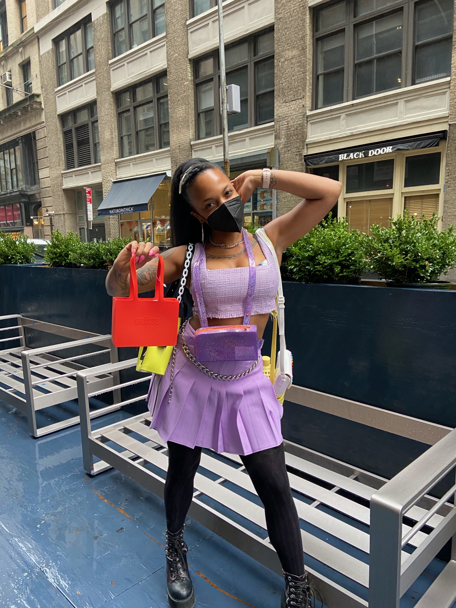 person poses in front of benches wearing lavender pleated skirt and matching top, holding 5 crossbody bags