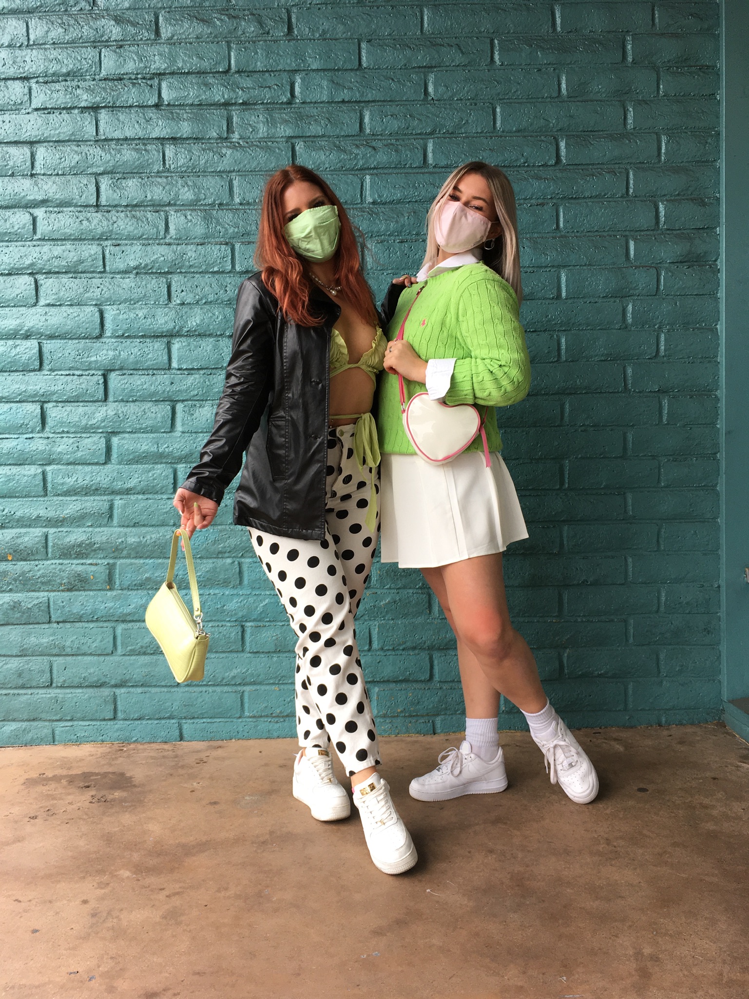Two people pose close together in front of teal brick wall. One person wears green bikini top underneath black leather jacket and polka-dot pants. The other wears a bright green Ralph Lauren sweater with white tennis skirt and heart-shaped bag