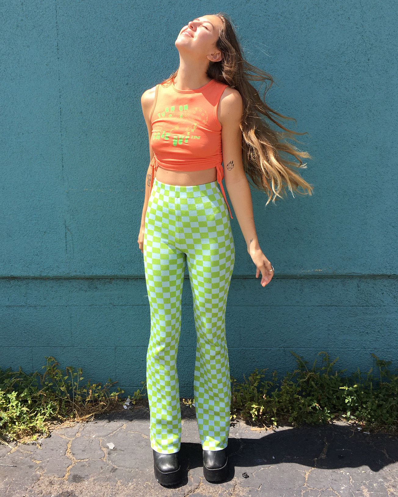 Person stands in front of teal wall with their face pointed towards the sun wearing cropped orange tank, green and white flares pants and black platform boots