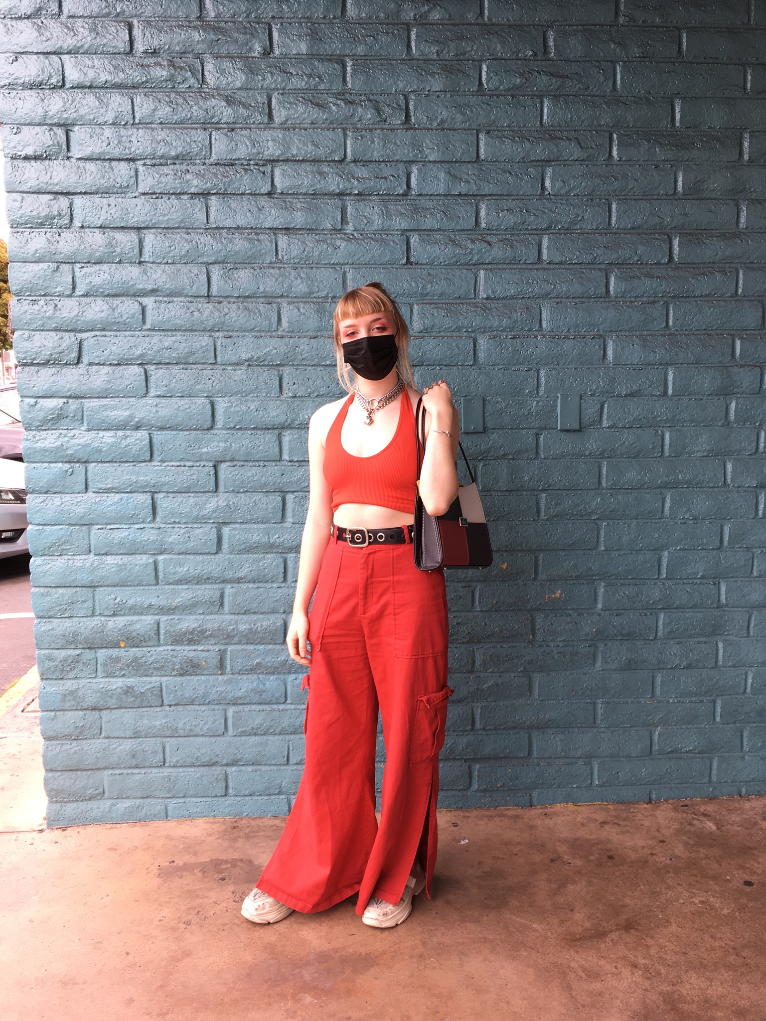 Person stands in front of teal wall wearing a black facemask, red halter top, silver chain necklaces, red cargo pants and patchwork leather shoulder bag