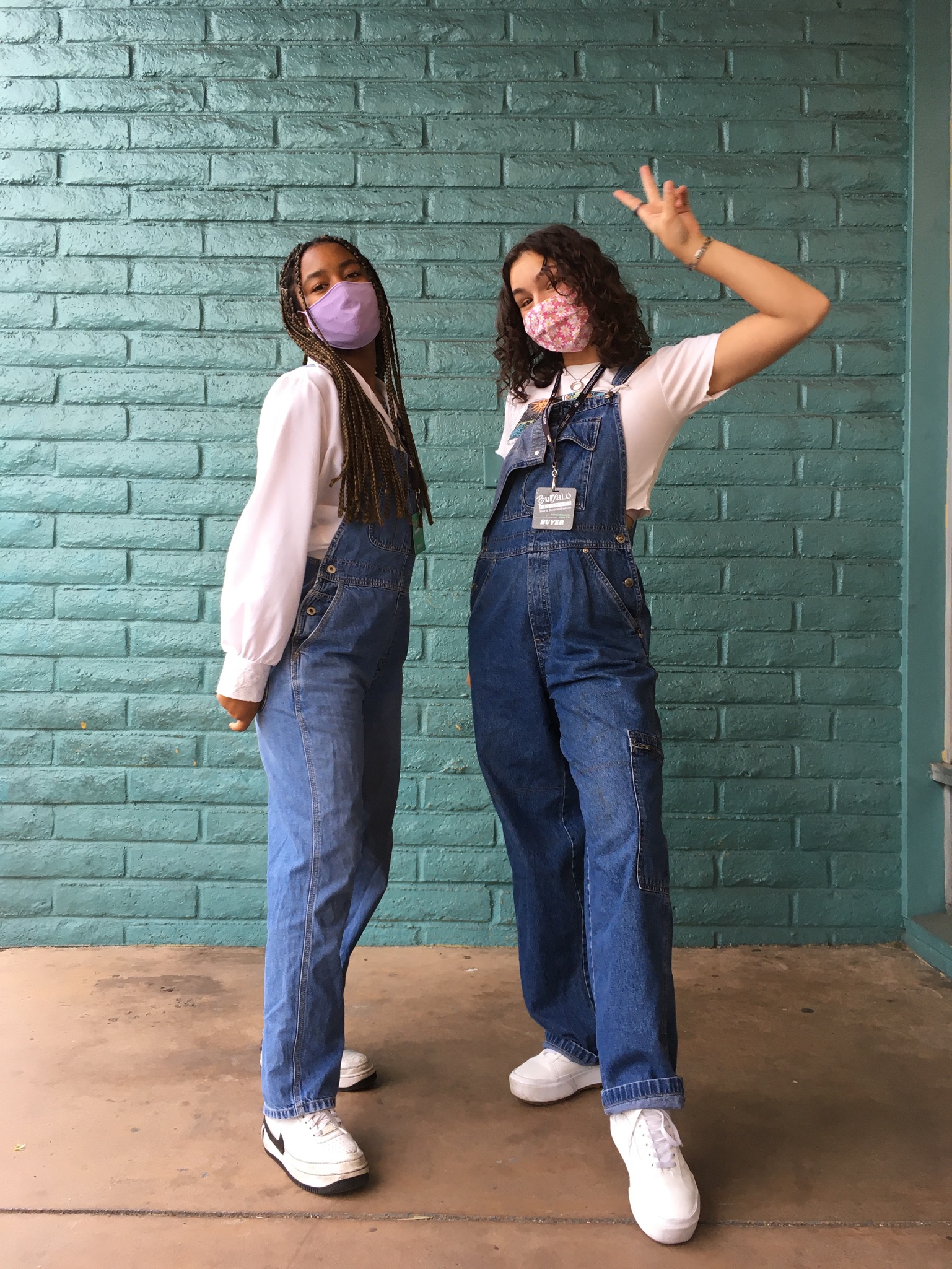 Two people stand posed in front of teal wall wearing denim overalls styled with white t-shirts and sneakers