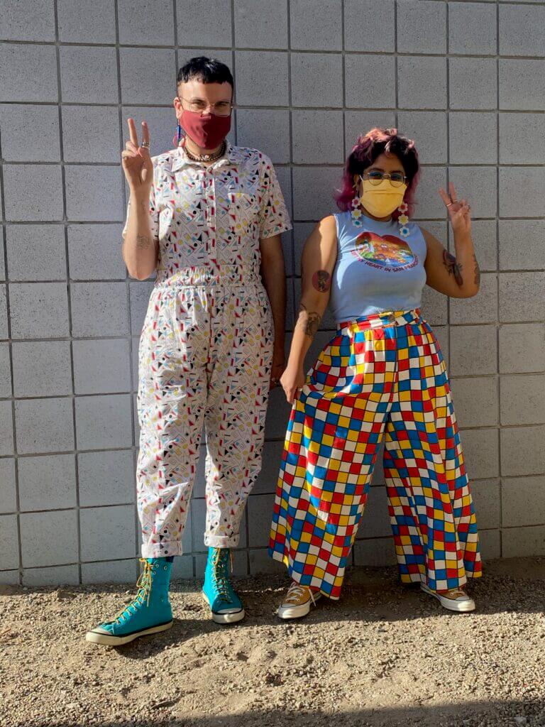 Two people holding up peace signs, one wearing an 80s-printed jumpsuit and the other primary-colored checkered pants and a grey tank top.