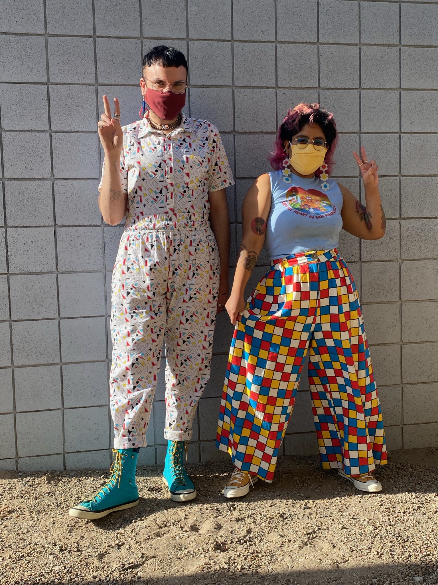 Two people holding up peace signs, one wearing an 80s-printed jumpsuit and the other primary-colored checkered pants and a grey tank top.