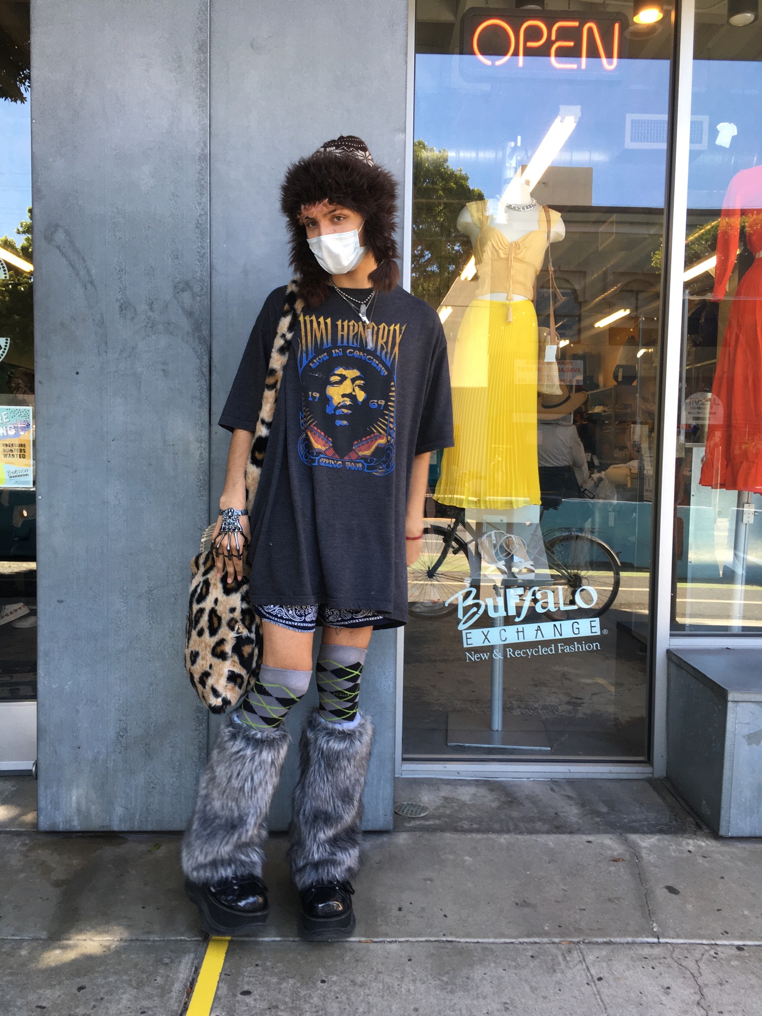 Person stands in front of 'open' sign at Buffalo Exchange Ventura wearing Jimi Hendrix t-shirt, bandana shorts, leopard print shoulder bagargyle knee socks and fuzzy leg warmers over black platform shoes.