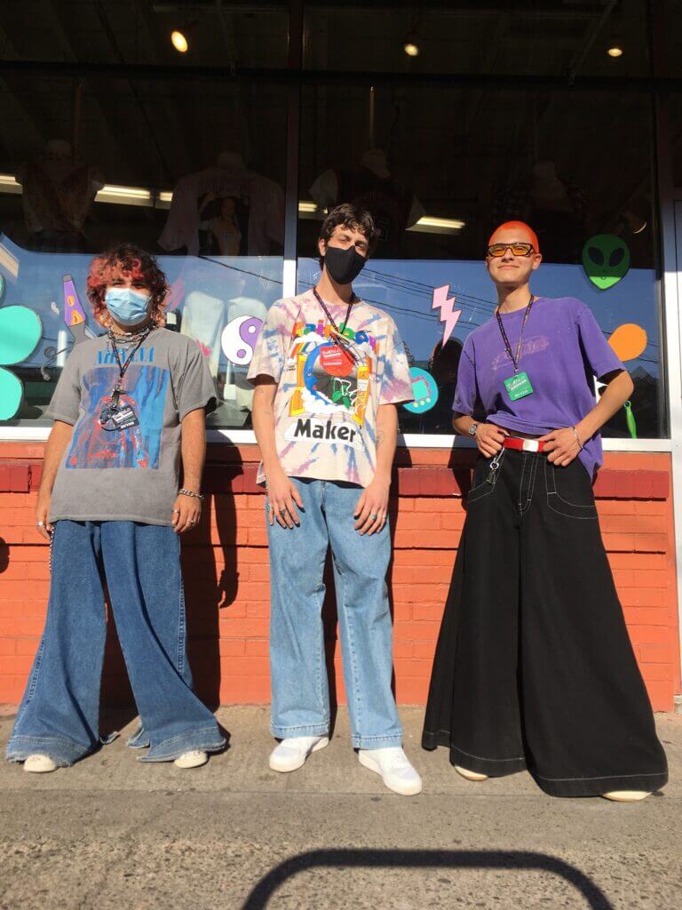 Three people stand in front of a window with 90s-inspired decals wearing graphic tees and oversized, wide leg denim