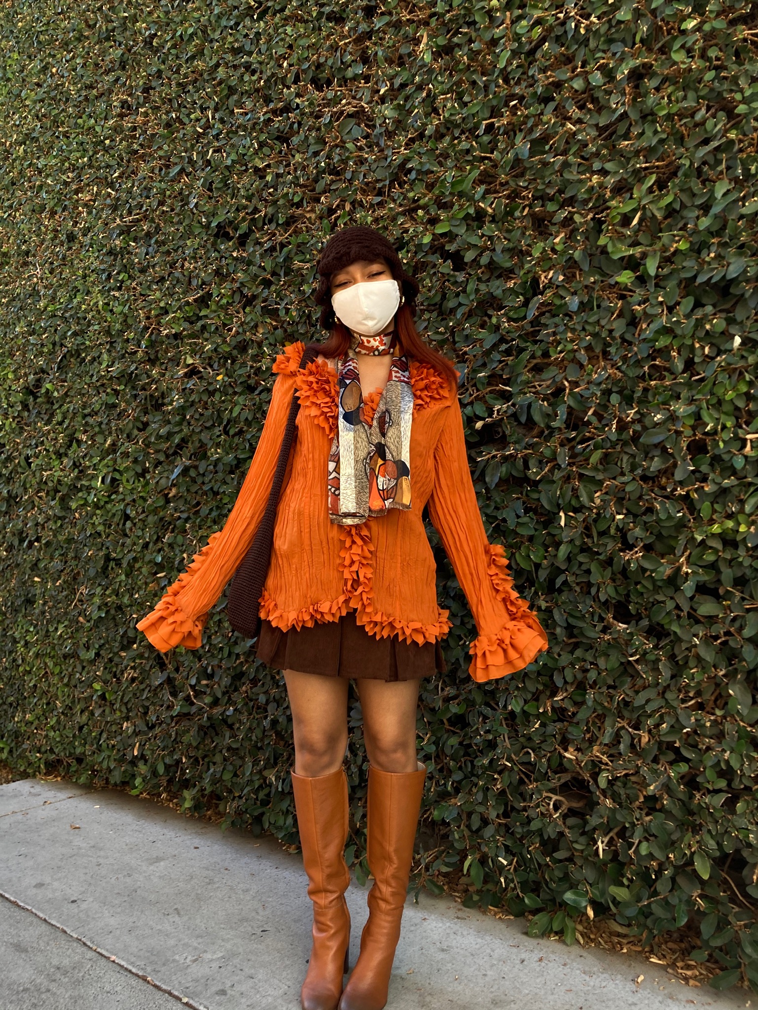 Person standing in front of shaped hedge wearing orange and brown-toned vintage inspired outfit with mini skirt and tall leather boots