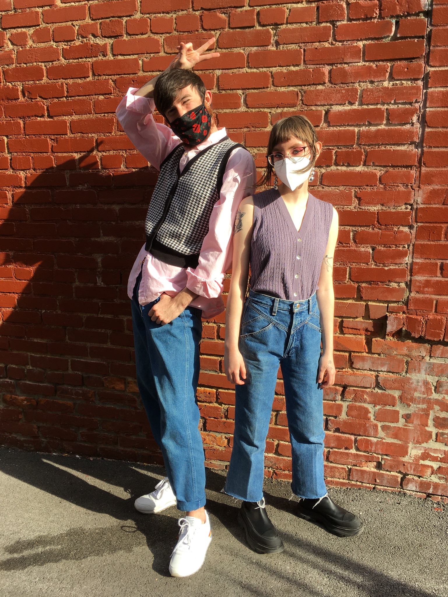 Two people stand posed in front of red brick wall. Left: wearing pink button-down shirt underneath houndstooth sweater vest, midwash denim and white sneakers. Right: wearing mauve knit sweater vest, midwash denim with western stitching and platform black boots