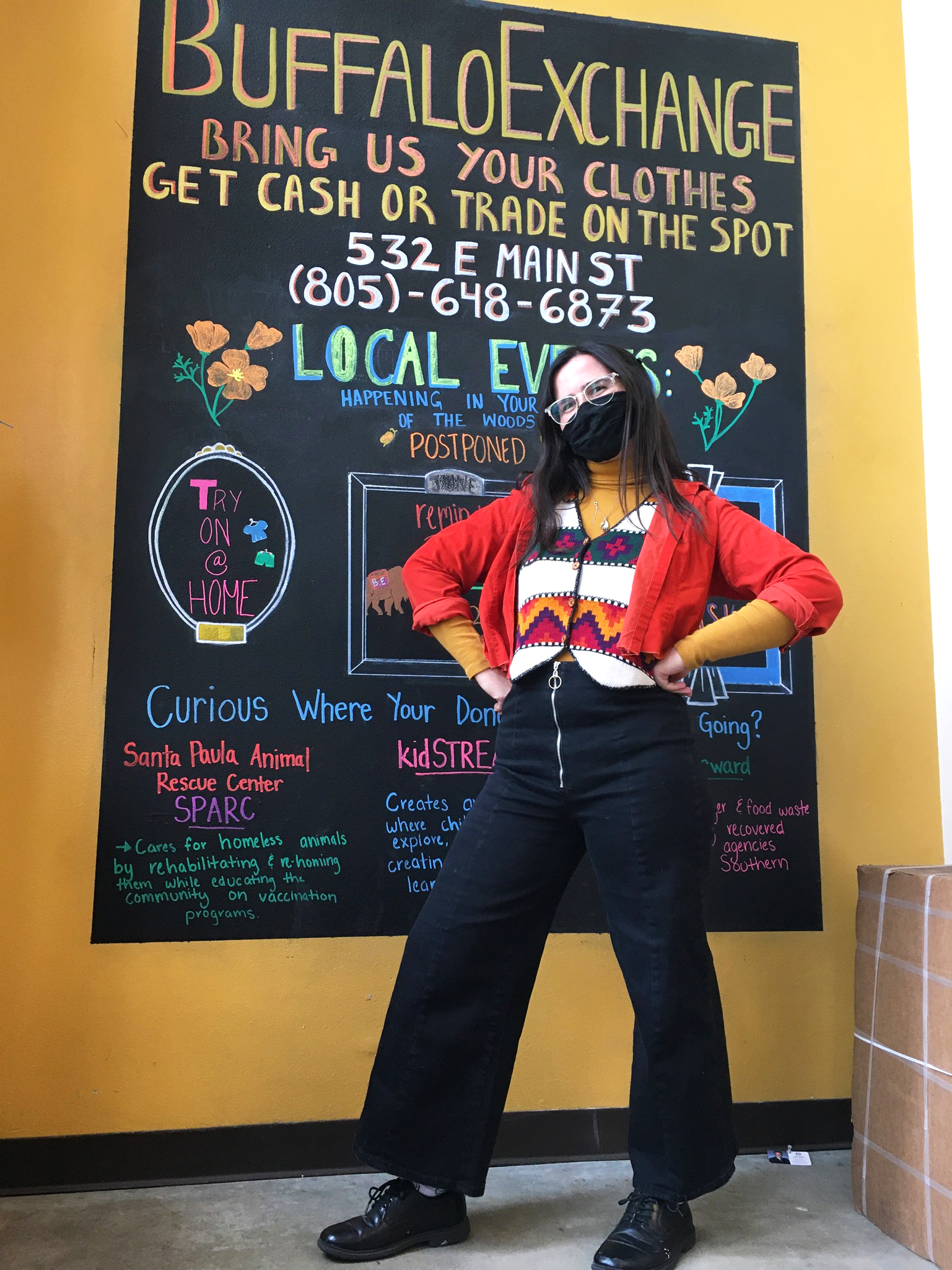 Buffalo Exchange employee in vest, layered orange sweaters and black pants stands in front of chalkboard painted on wall