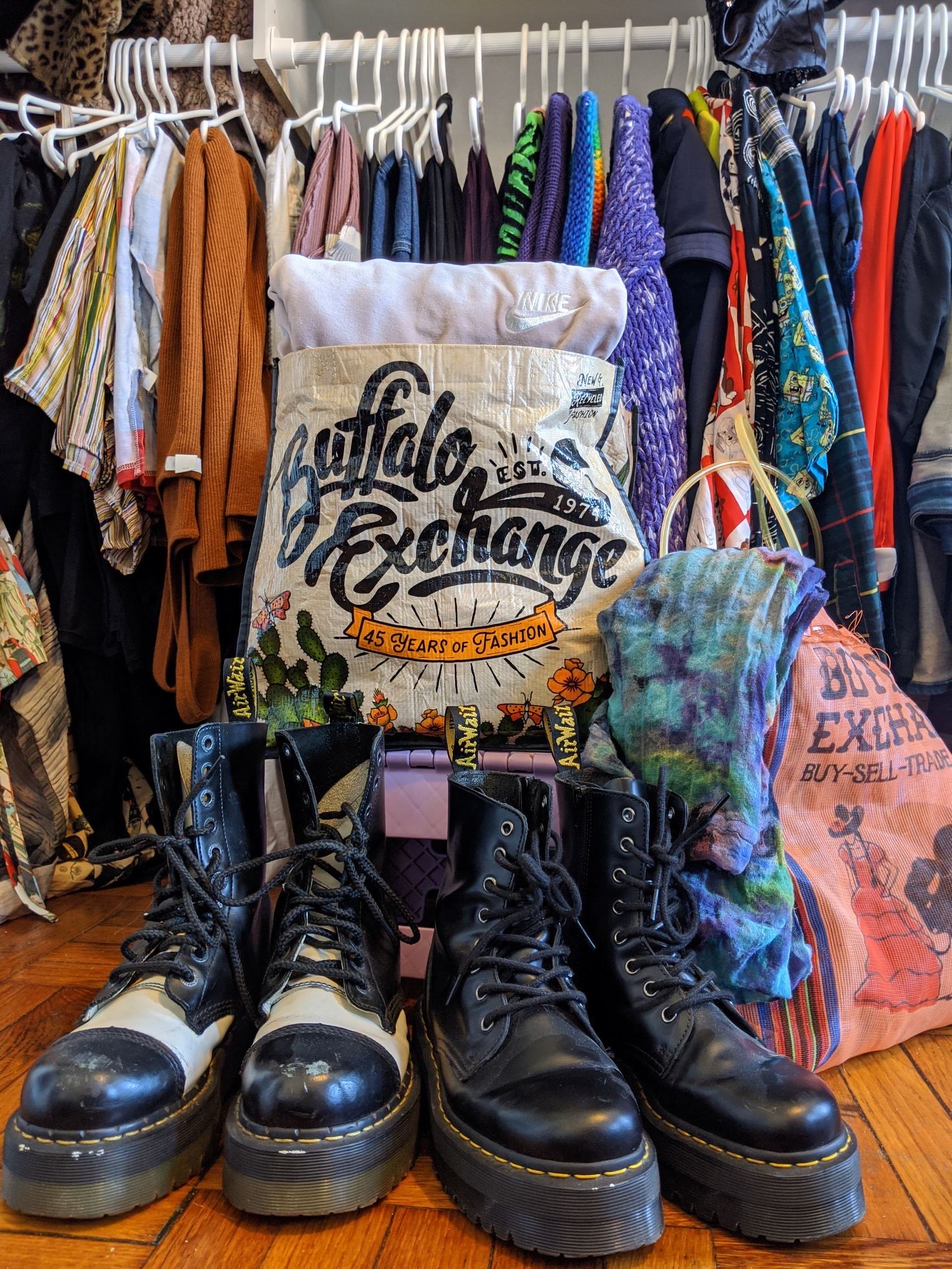 Two pairs of platform Dr.Martens boots front of Buffalo Exchange tote bags and a rack full of clothing