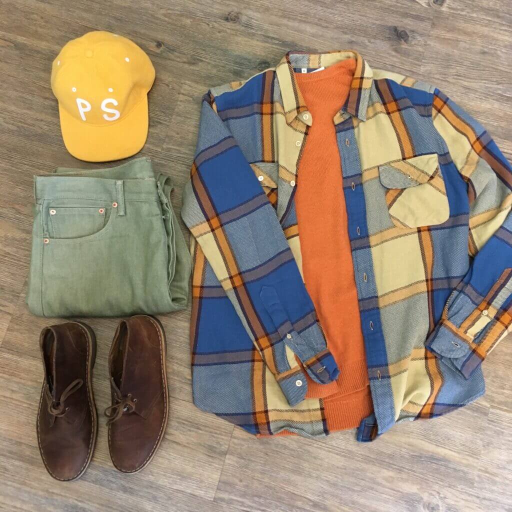 Flat lay of earth tone clothing including yellow ballcap, green jeans, brown suede boots, orange sweater and blue and orange plaid shirt