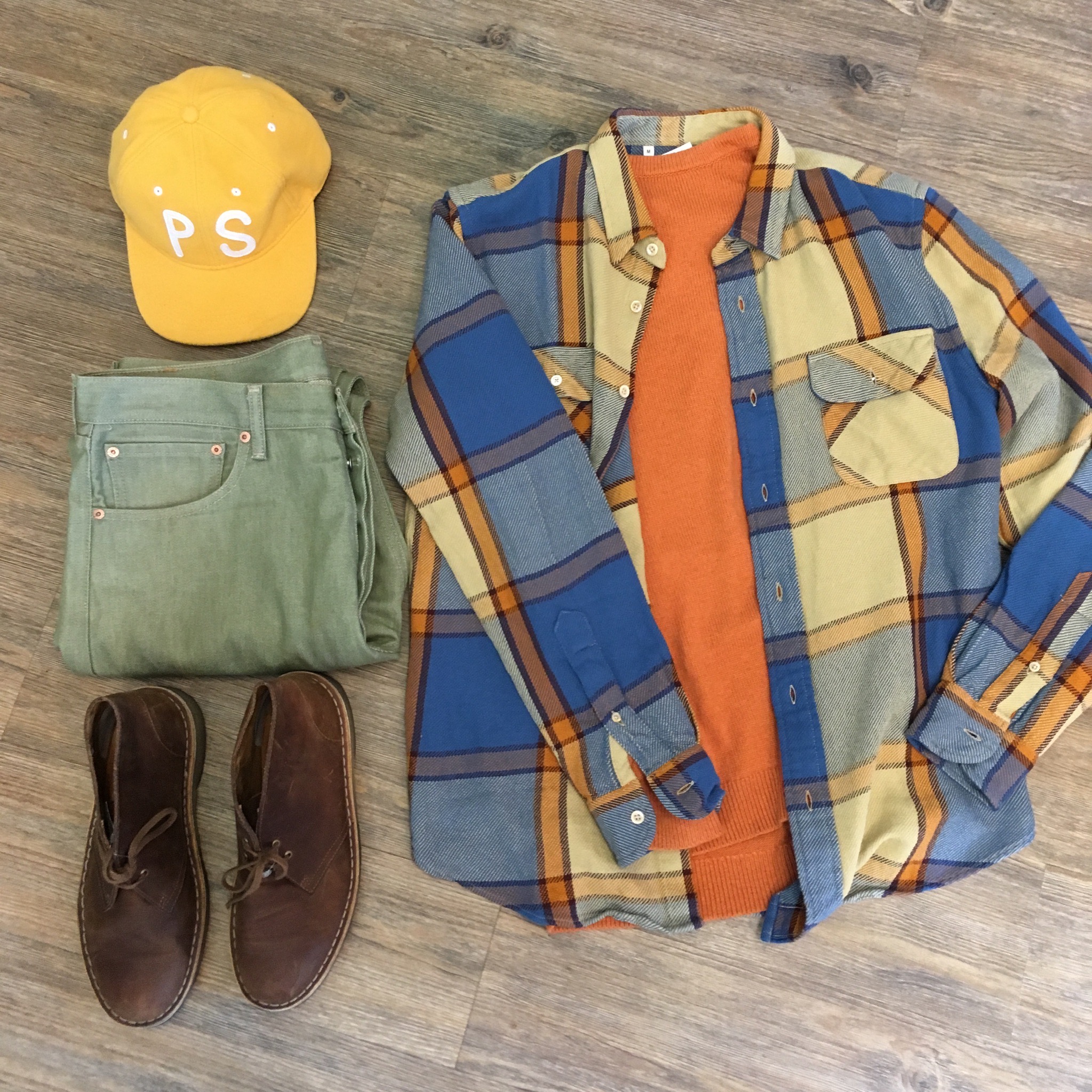 Flat lay of earth tone clothing including yellow ballcap, green jeans, brown suede boots, orange sweater and blue and orange plaid shirt