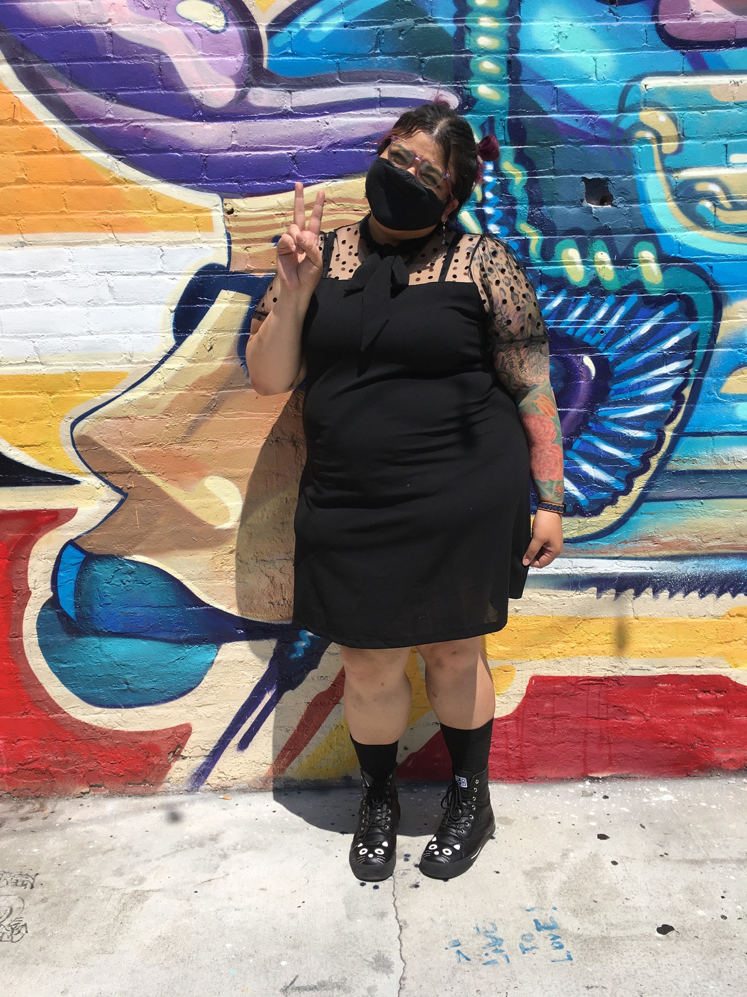 person standing in front of colorful mural wearing all black outfit and giving the peace sign