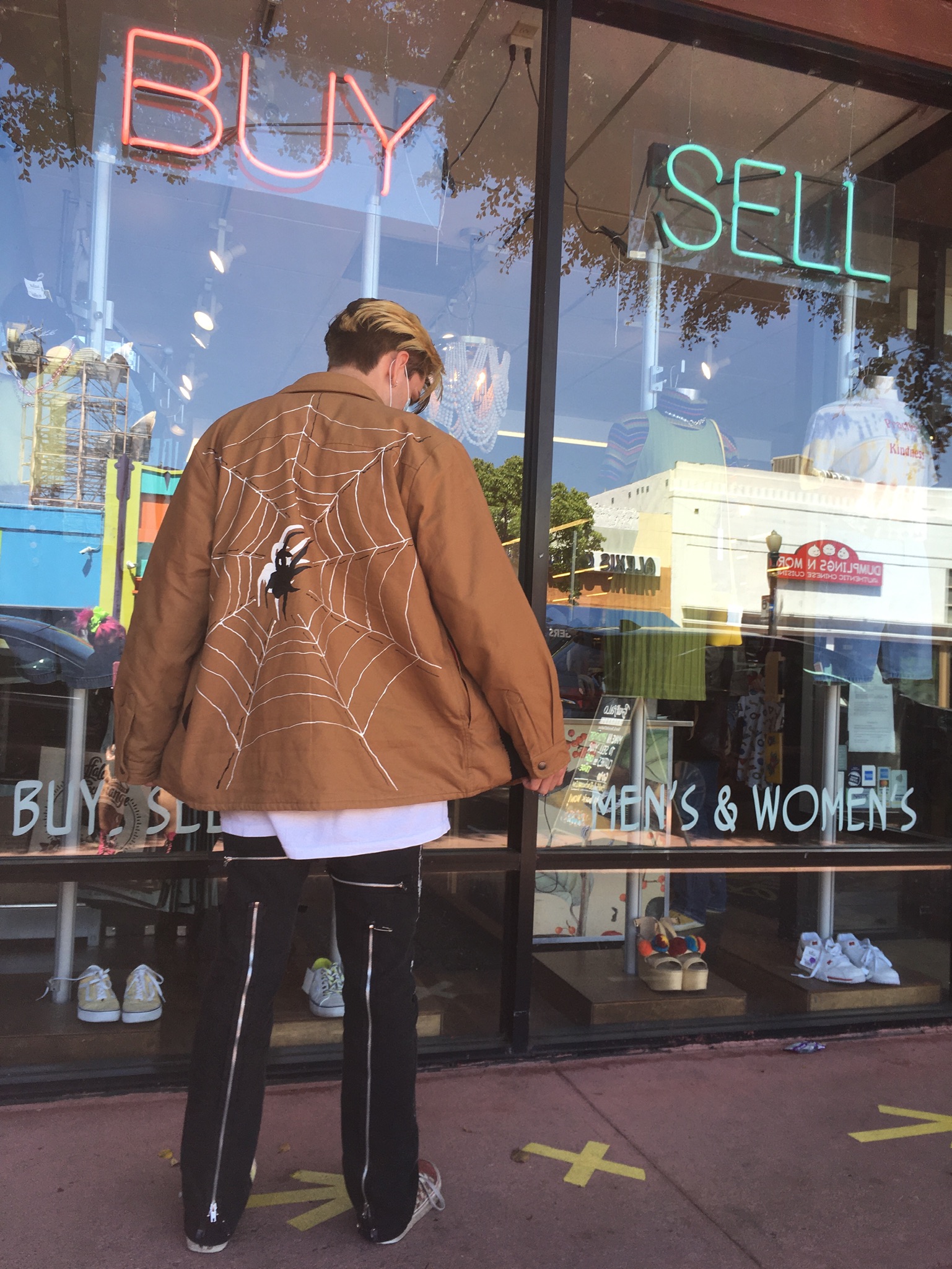 Person standing beneath neon signs reading Buy and Sell with back turned showing hand-painted spider web on brown jacket