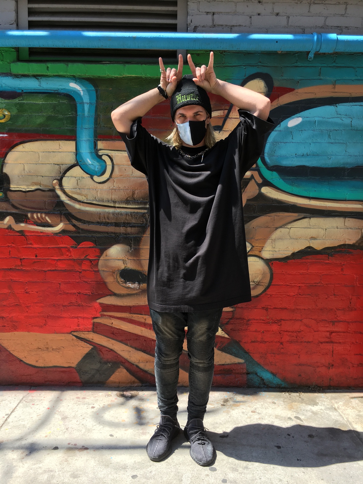 Buffalo Exchange Ventura Person standing in front of mural wearing oversized black shirt, black beanie and black and white face mask
