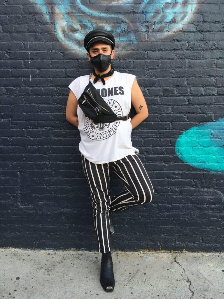 Person standing in front of black brick wall wearing black boots, black and white striped pants, Ramones tee and black accessories