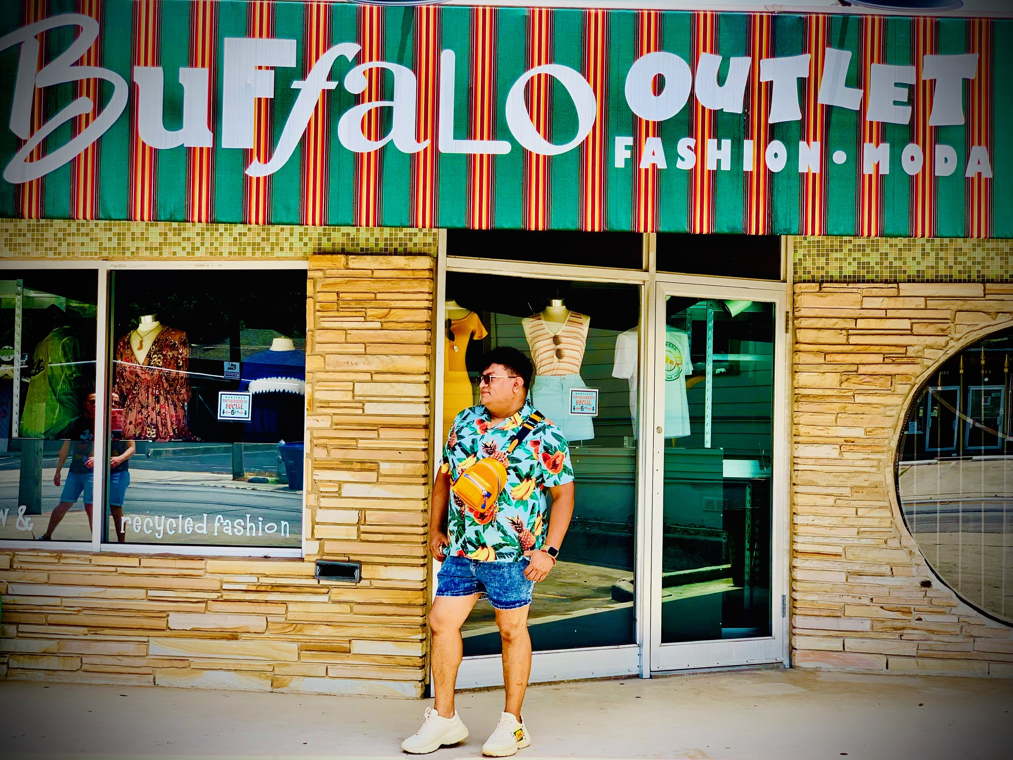 Felix in Hawaiian shirt, jean shorts and yellow crossbody bag in front of Buffalo Outlet store
