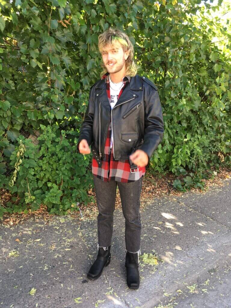 Person standing outside in front of vines wearing black boots, red plaid shirt and leather jacket