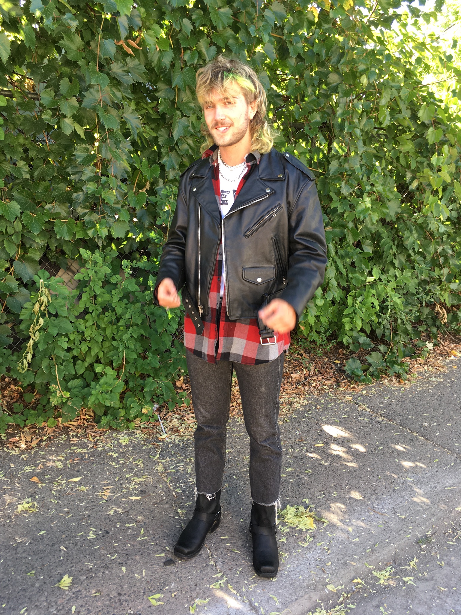Person standing outside in front of vines wearing black boots, red plaid shirt and leather jacket