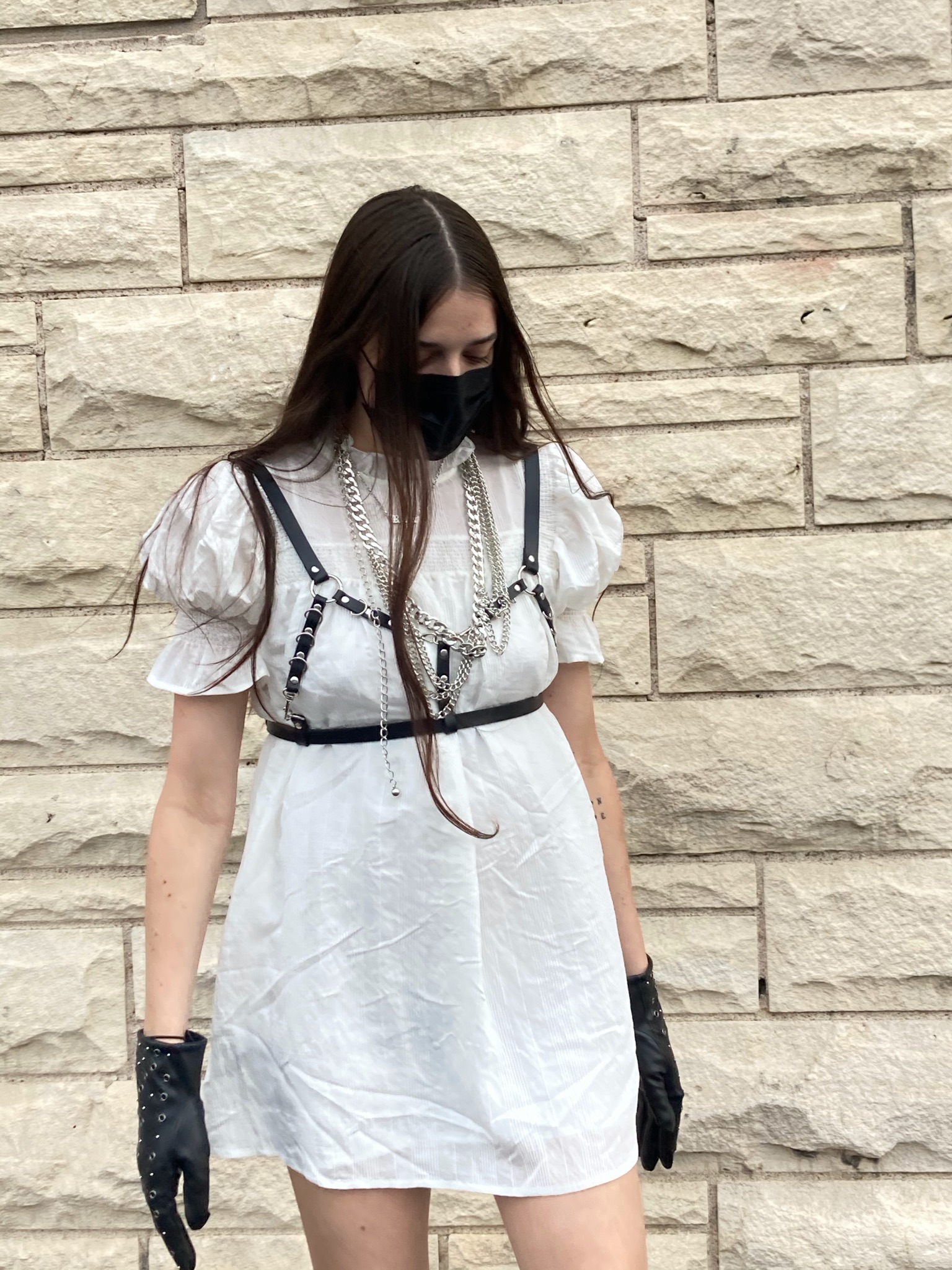 Person in front of cream brick wall wearing white dress with black harness and gloves