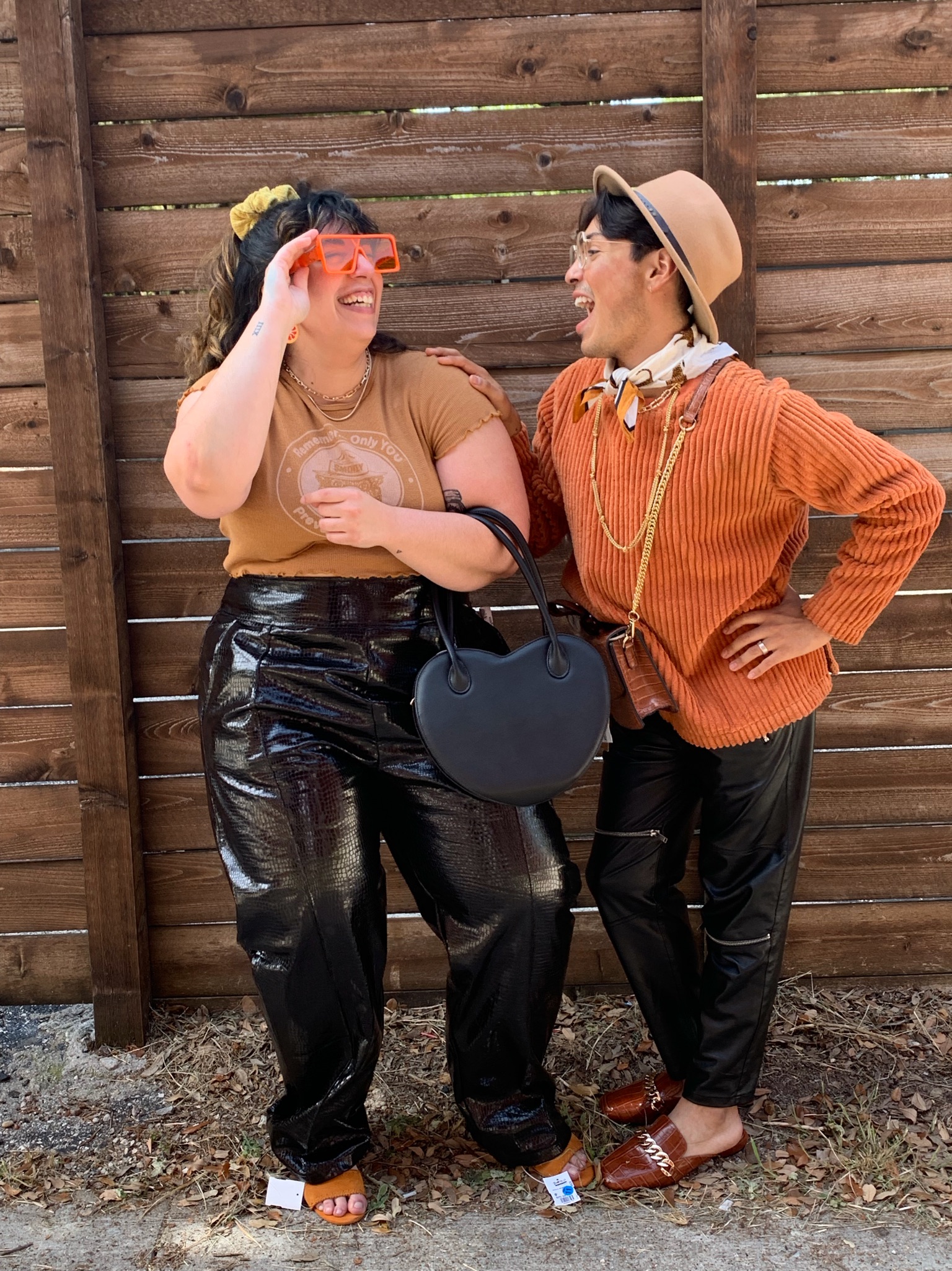 2 people in front of a wood fence laughing and wearing orange tops and black pleather pants