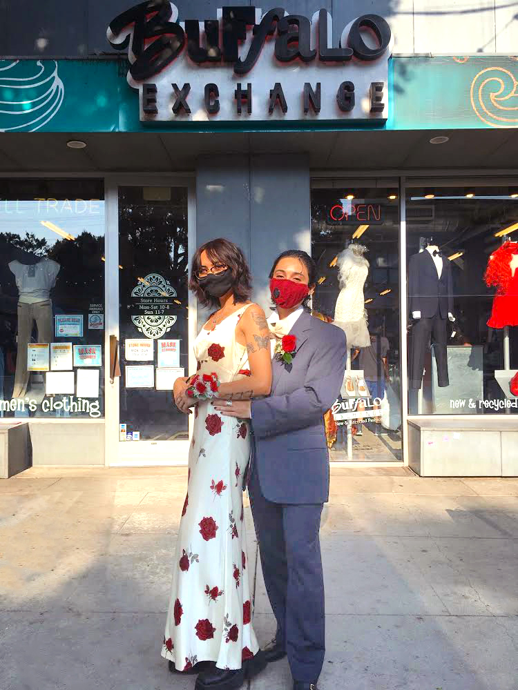 2 People dressed in prom outfits in front of Buffalo Exchange Ventura, one wearing rose print white dress and one wearing blue/gray suit with rose corsage