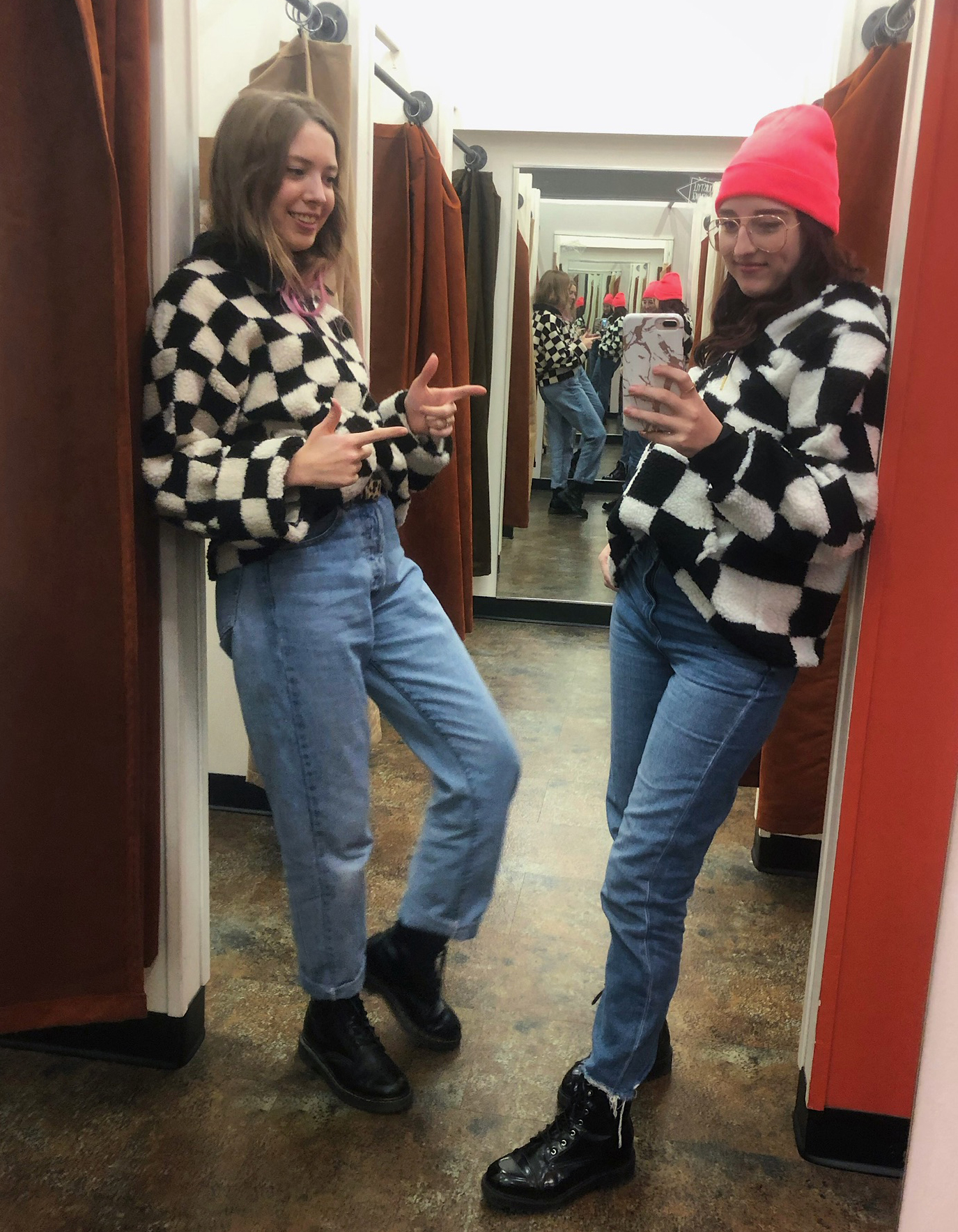 two people posing in a dressing room wearing matching checkered printed sweaters, blue jeans and black boots