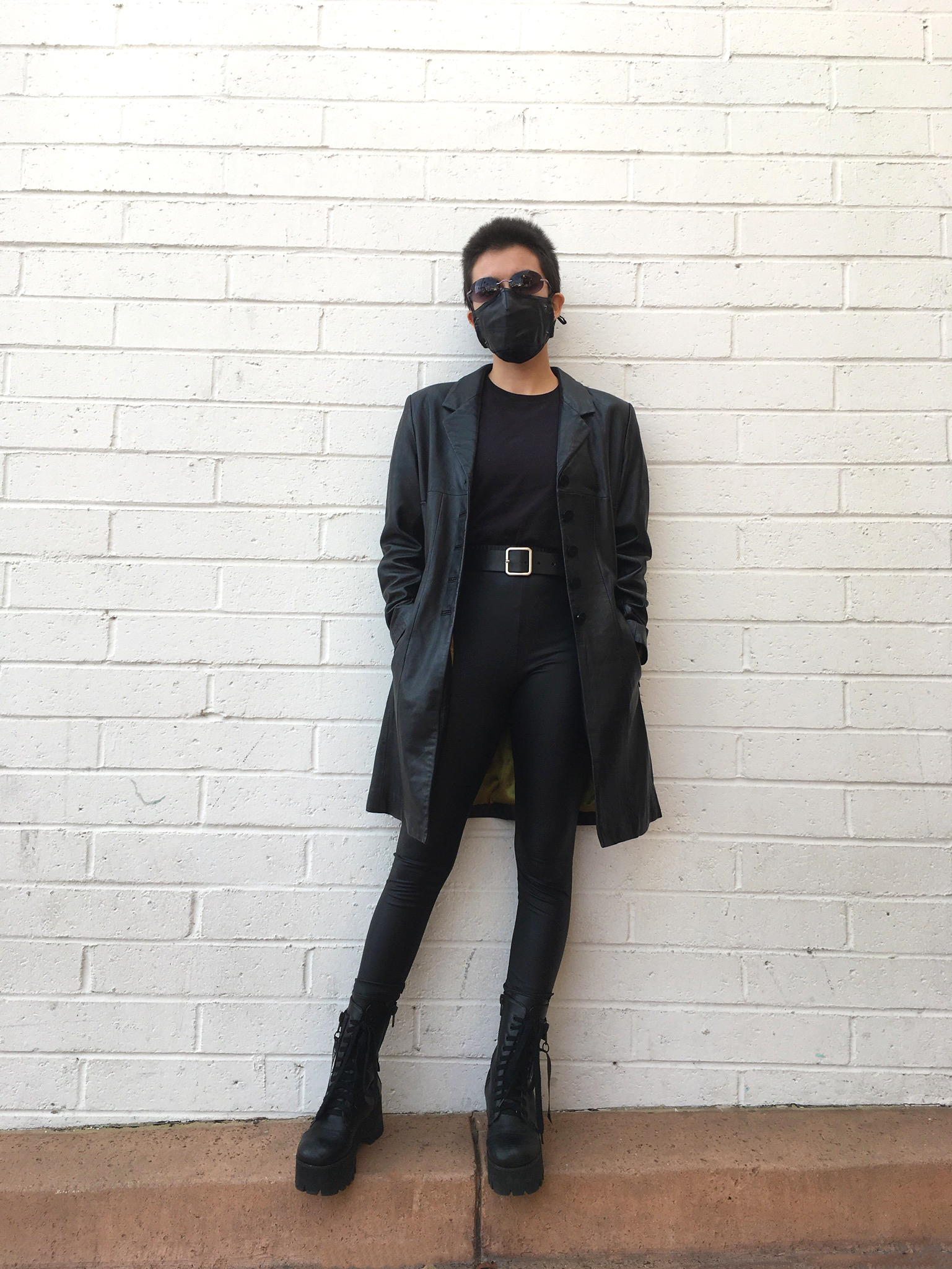 A person standing again a white brick wall wearing a full black outfit with a pleather trench coat, black skinny jeans and platform boots