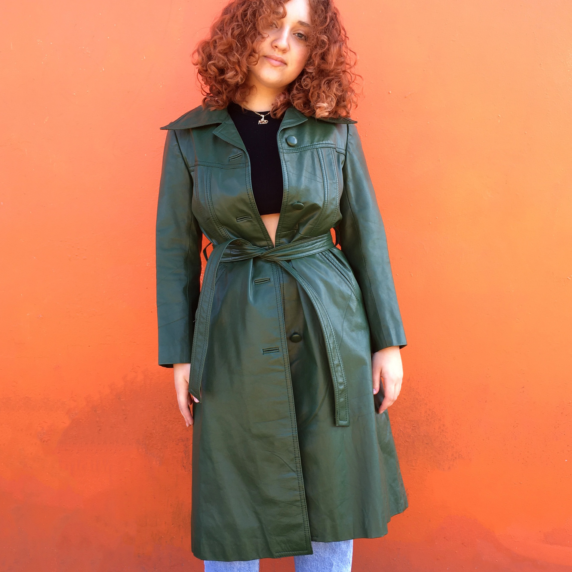 A person with curly red hair standing against an orange wall wearing a vintage green leather coat and black crop top