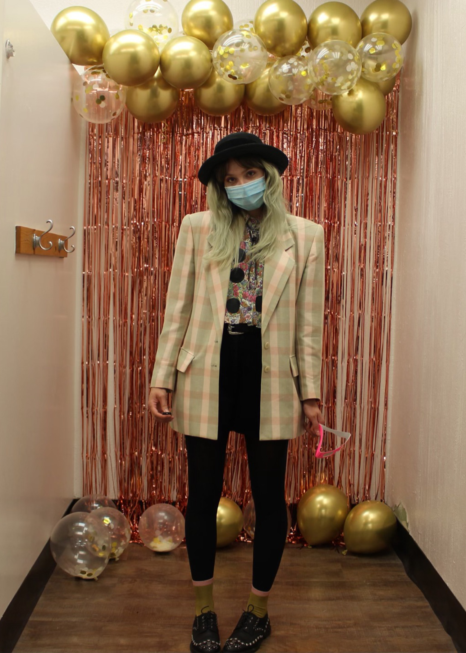 A person wearing an oversized pastel plaid jacket and a bowler hat with balloons in background
