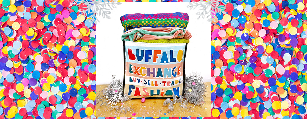 Buffalo Exchange tote bag that reads "Buffalo Exchange, Buy Sell Trade Fashion" is stacked with clothing and sits on a light wood table surrounded by confetti and silver tinsel. The background is a close-up of rainbow confetti