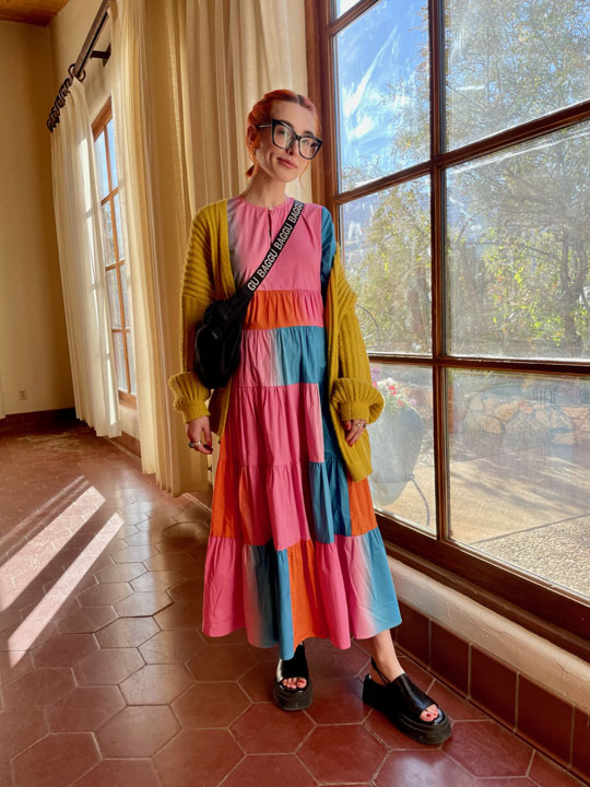 a woman posing by a window wearing a patch work colorful dress and a yellow cardigan