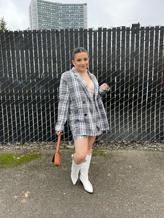 a woman posing by a fence wearing a grey plaid jacket and matching mini skirt, white boots and a brown