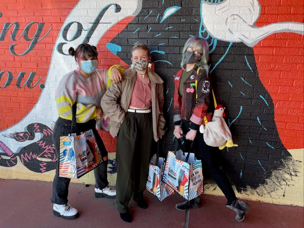 Three shoppers standing next to mural while holding Buffalo Exchange tote bags